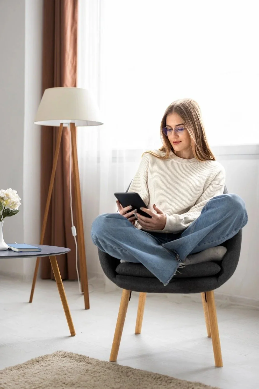 A young woman with glasses, sitting cross-legged in a black chair, is looking at her phone in a bright room with a floor lamp, a small table with a vase of flowers, and curtains.