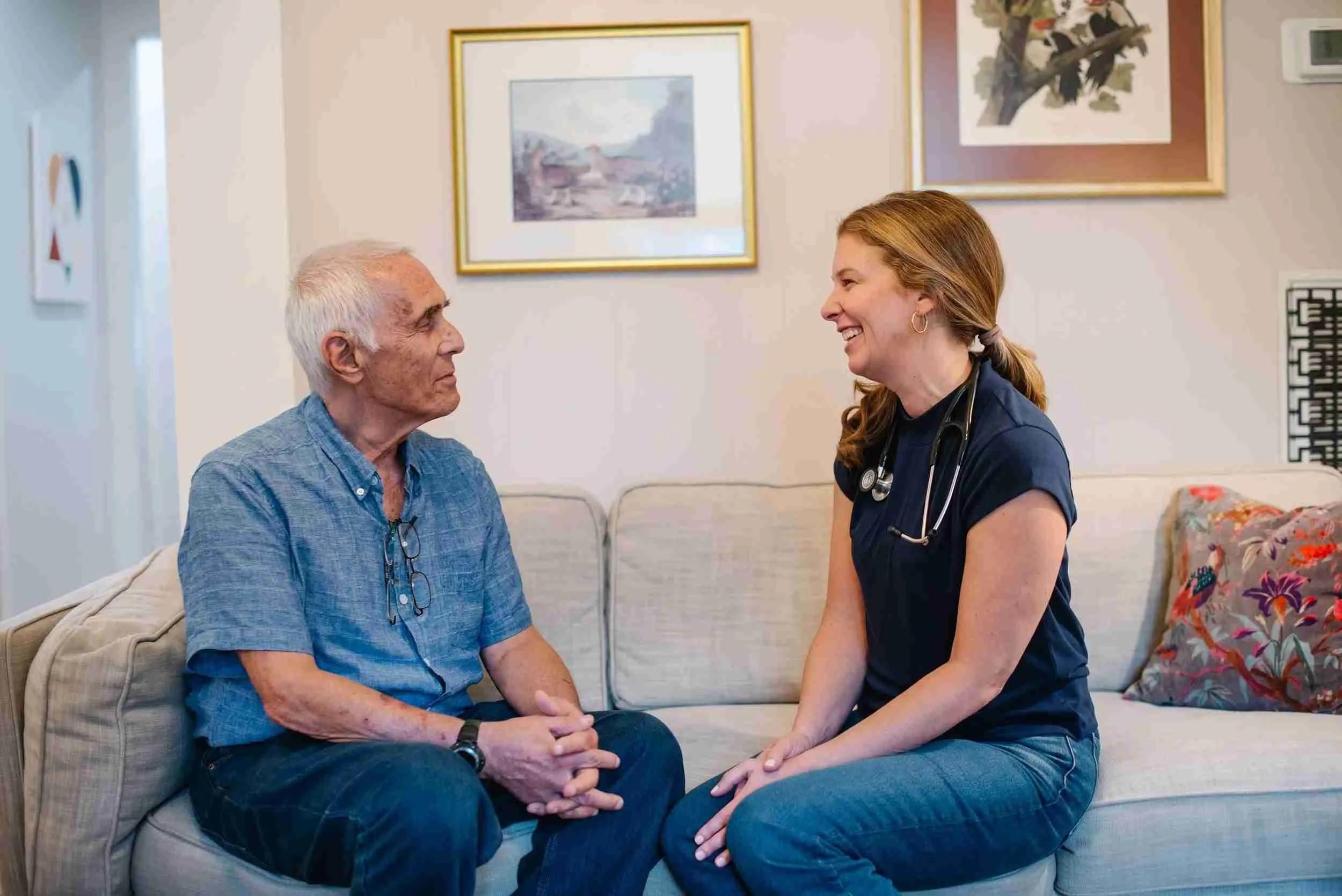 A healthcare professional, a woman with red hair and wearing navy scrubs with a stethoscope around her neck, is sitting on a beige couch and smiling at an elderly man with white hair and a blue shirt. They are engaging in a friendly conversation in a living room with framed artwork on the wall.