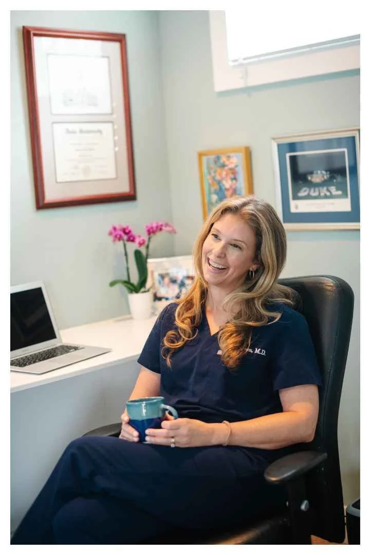 A woman in medical scrubs sitting in an office, holding a mug, smiling. There are framed certificates and artwork on the wall, a laptop on the desk, and a potted orchid plant.
