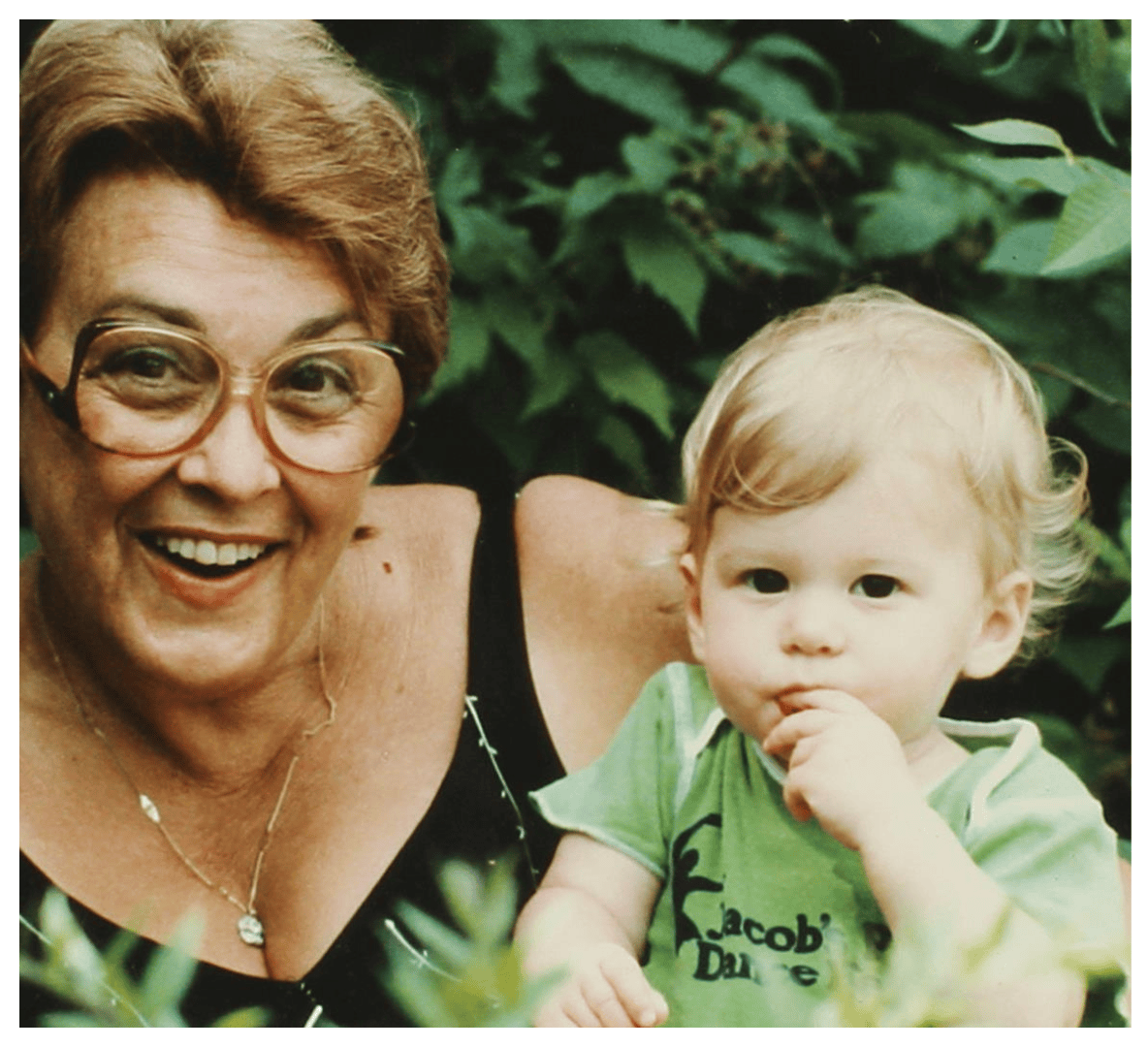 A happy woman with glasses and a necklace smiling with a young child with blonde hair and a green shirt, surrounded by green foliage.