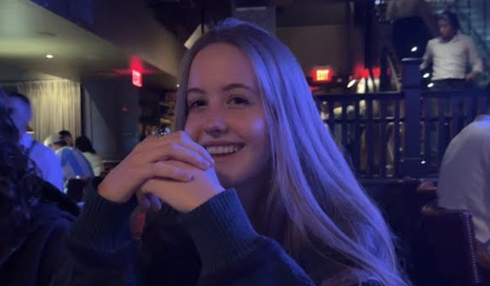 A young woman with long blonde hair smiling and resting her chin on her hands in a dimly lit indoor setting, likely a restaurant or bar, with people and a staircase visible in the background.