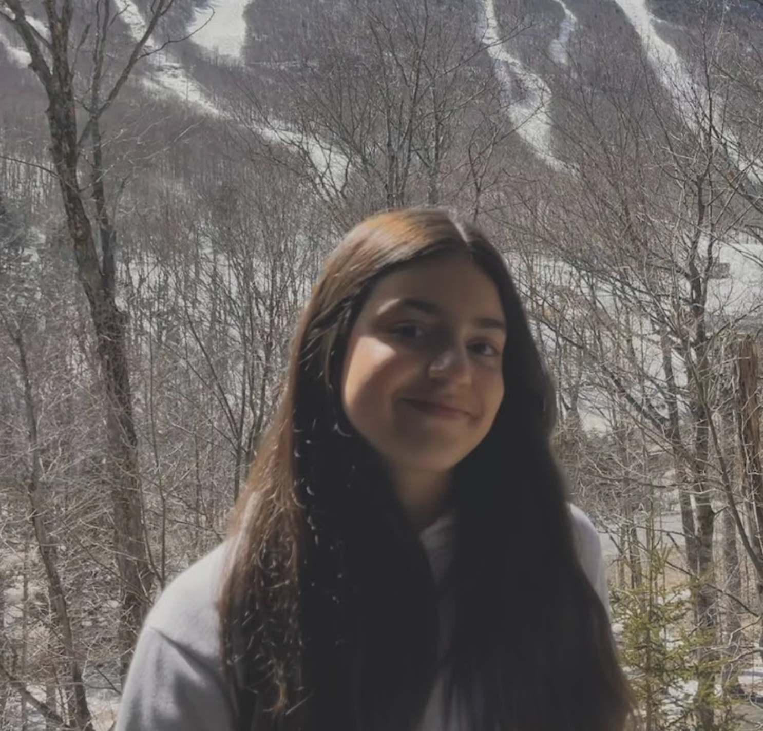 A young woman with long dark hair stands outside against a winter landscape with snow-covered trees and mountains in the background, smiling softly at the camera.