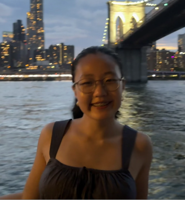 A woman with glasses smiling near the water on a boat at dusk, with a city skyline and a bridge illuminated in the background.