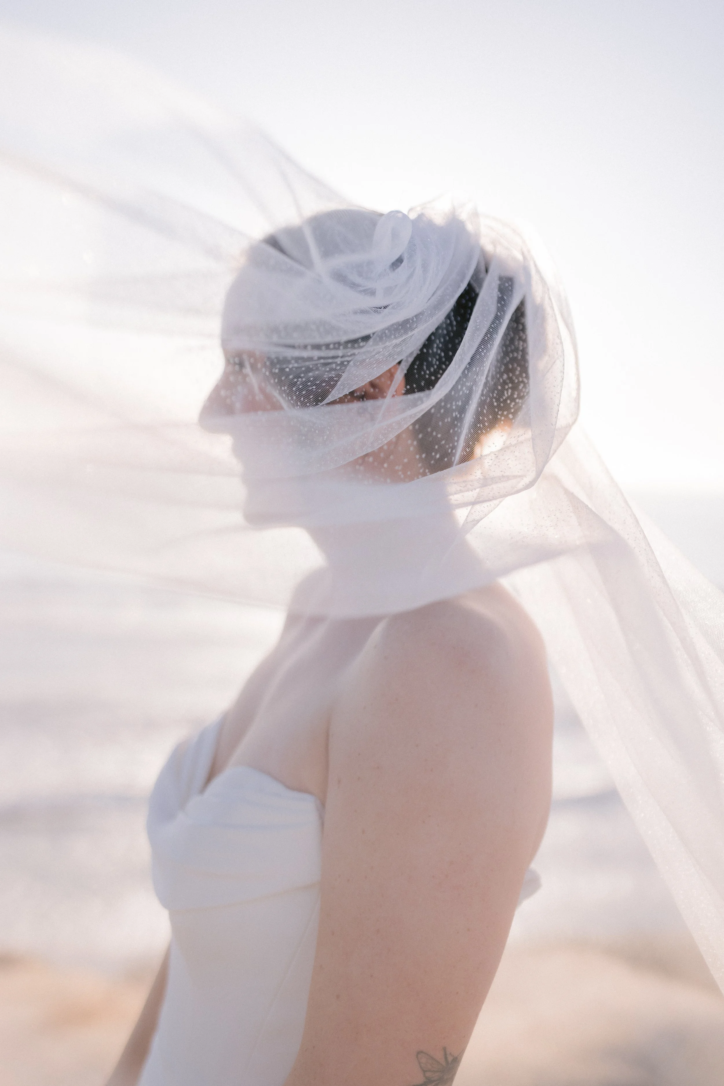 A woman wearing a white wedding dress and a sheer veil with small dots, standing outdoors with beach and ocean in the background.