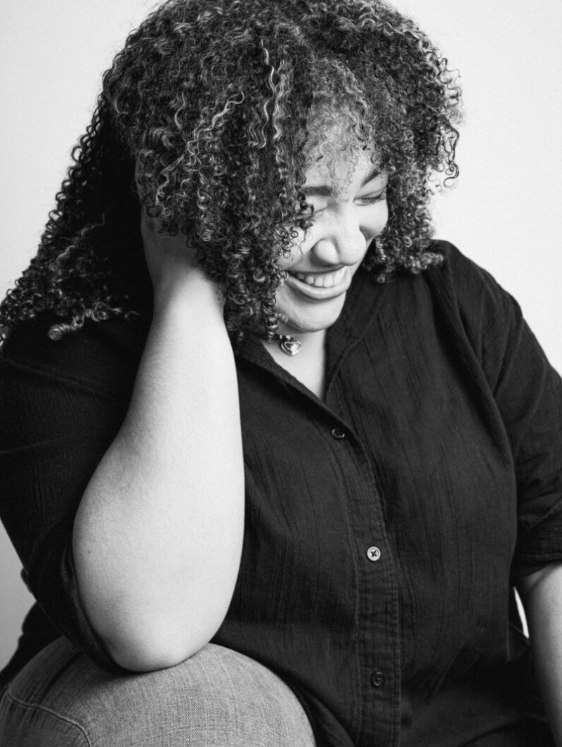 A woman with curly hair sitting with her head resting on her hand, smiling with her eyes closed, in black and white.