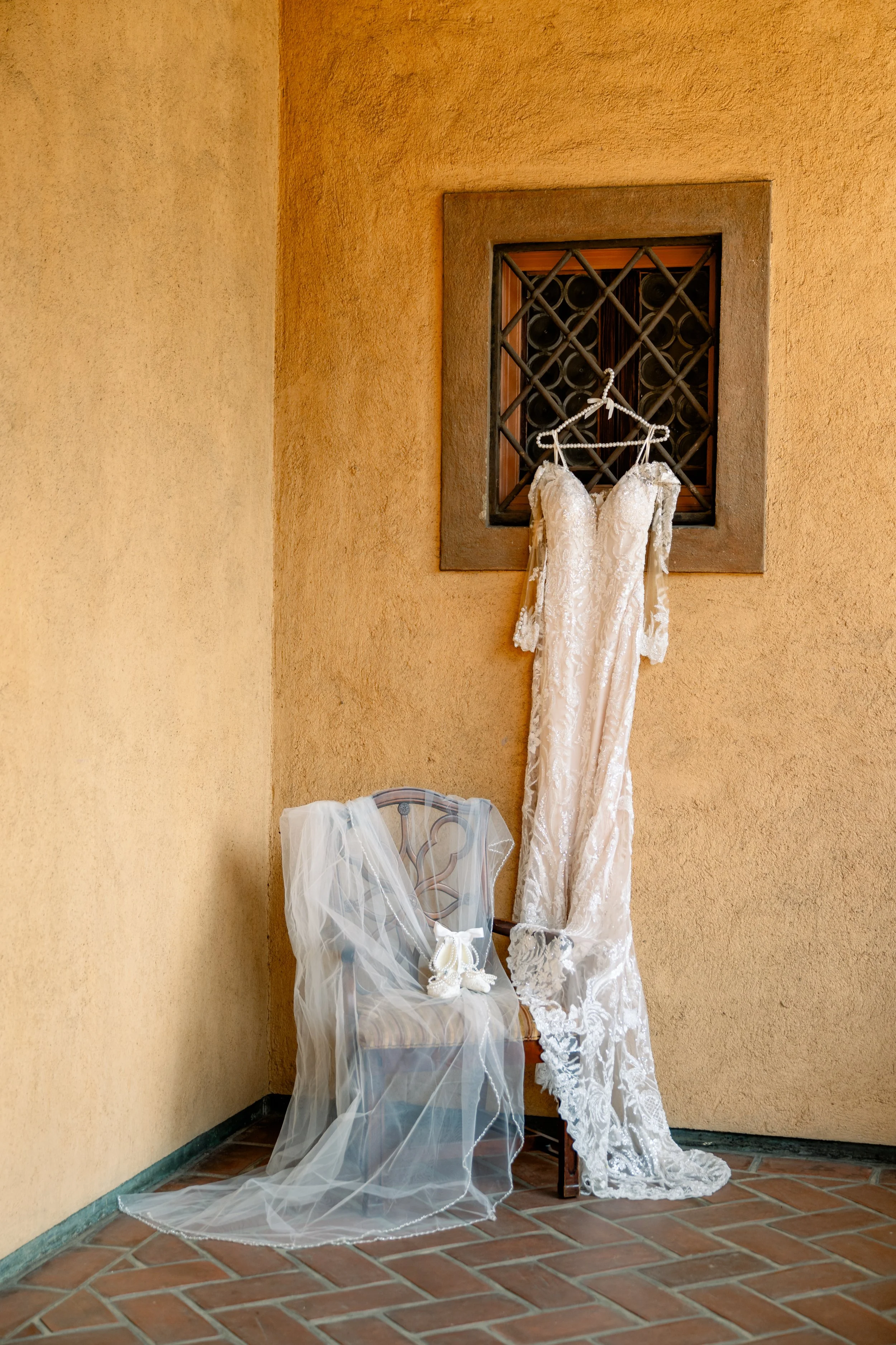 A wedding dress hangs on a decorative hanger on a small window with wrought iron bars, beside a chair draped with sheer fabric and a small bouquet on it, set against a yellow stucco wall.