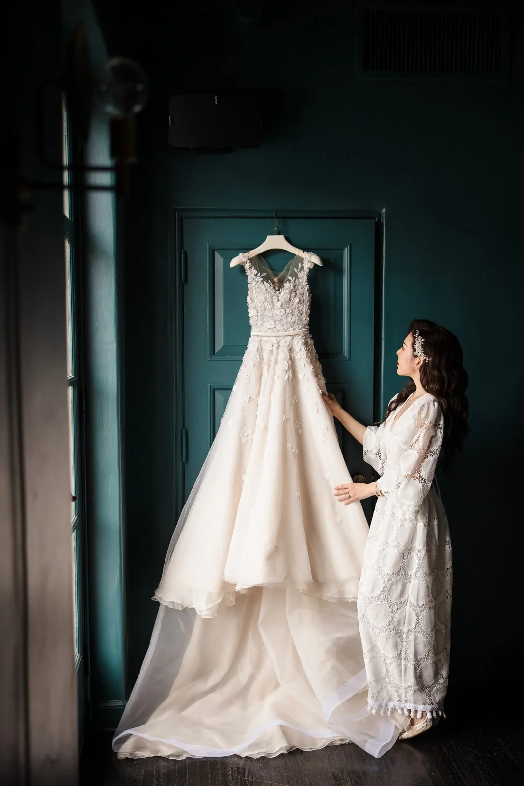 A woman in a white lace dress looking at a wedding gown hanging on a door. The gown is detailed with lace and floral embellishments, and the woman is adjusting or examining it in a room with dark walls.