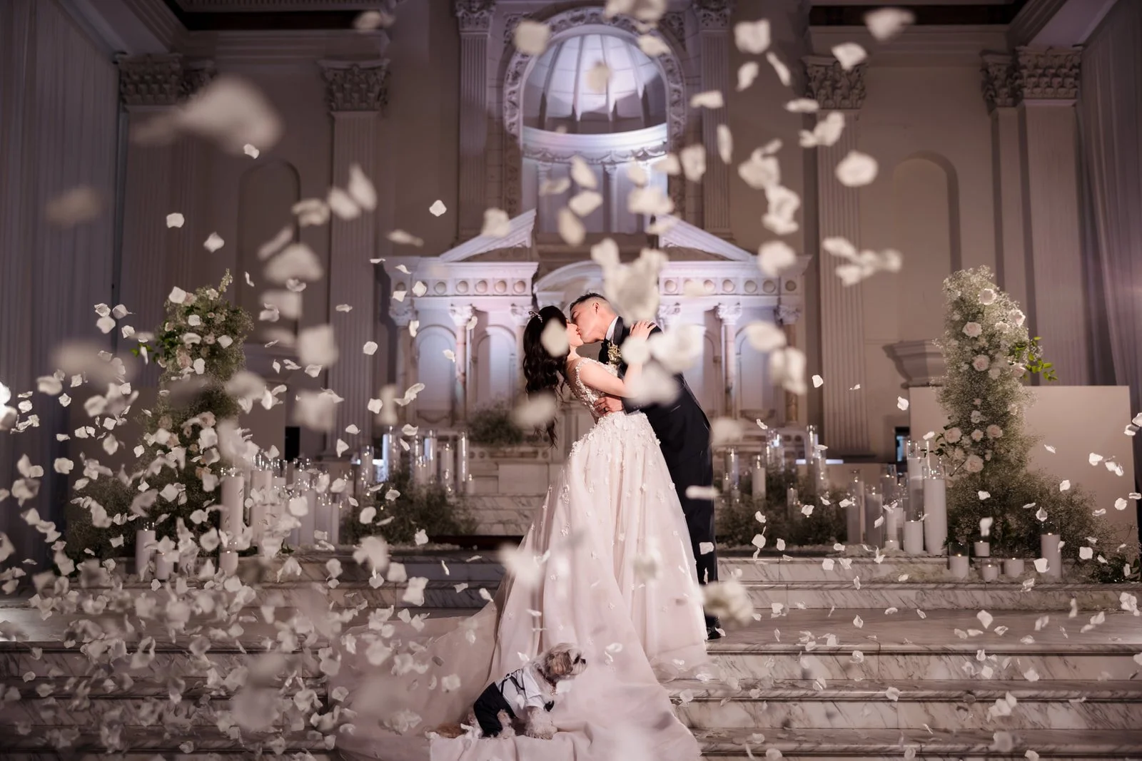 Bride and groom kissing on wedding altar with rose petals falling, wedding dress, tuxedo, church setting, and small dog in tuxedo