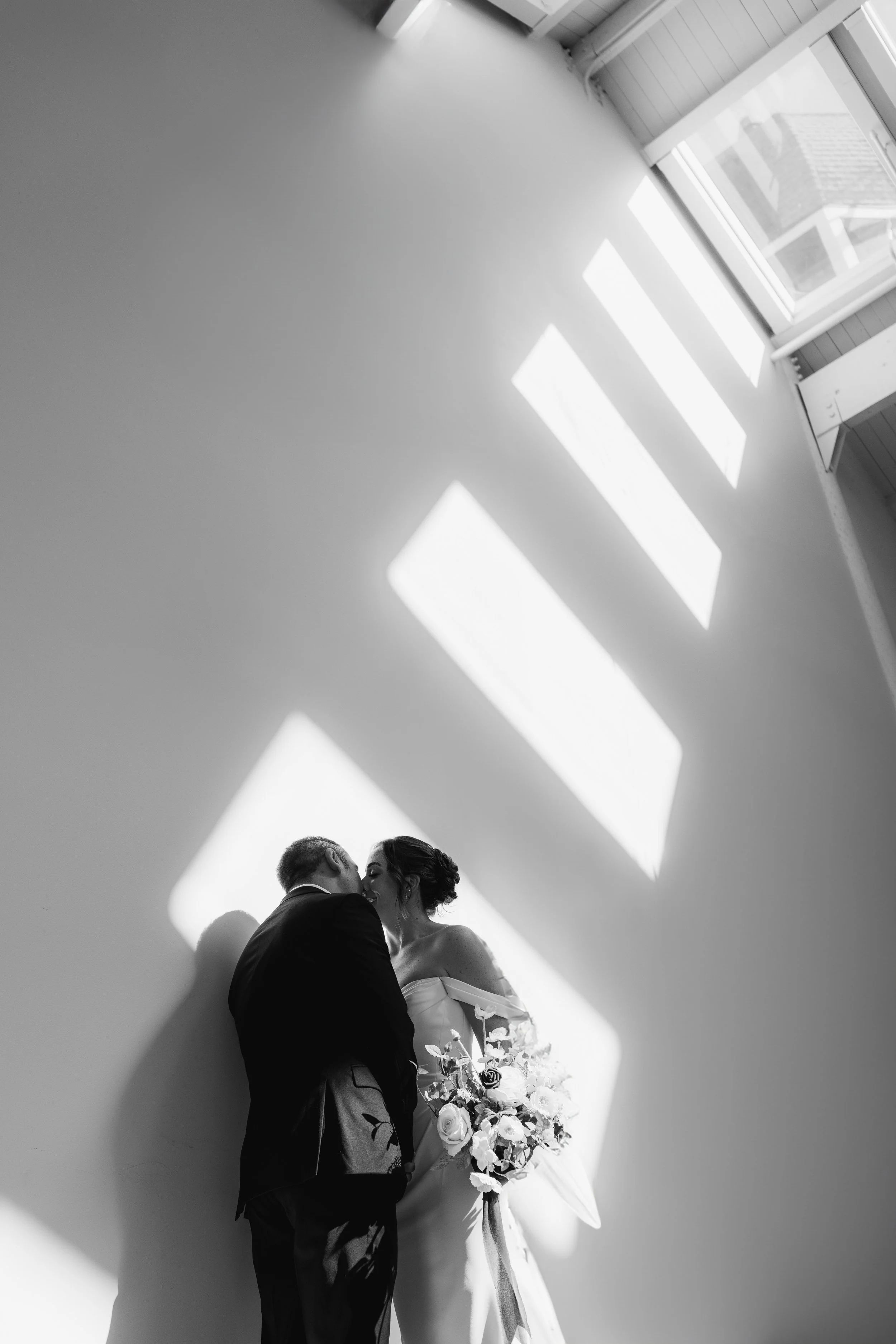 A black and white photo of a bride and groom sharing a kiss inside a room with sunlight streaming through large windows, casting rectangular shadows on the wall behind them. The bride is holding a bouquet of flowers.