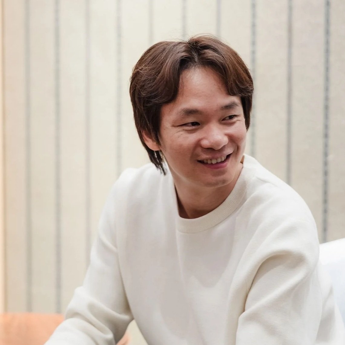 A smiling young man with medium-length brown hair wearing a white sweatshirt, sitting indoors against a ribbed wall backdrop.