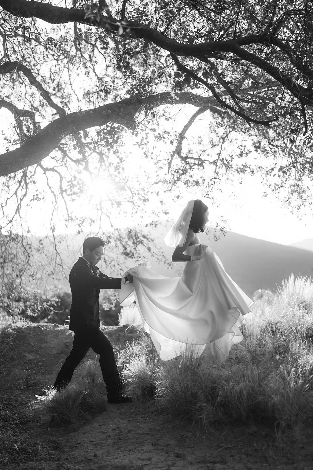A couple in wedding attire outdoors, with the man helping the woman in a flowing gown to step up on a natural trail under trees with sunlight streaming through.