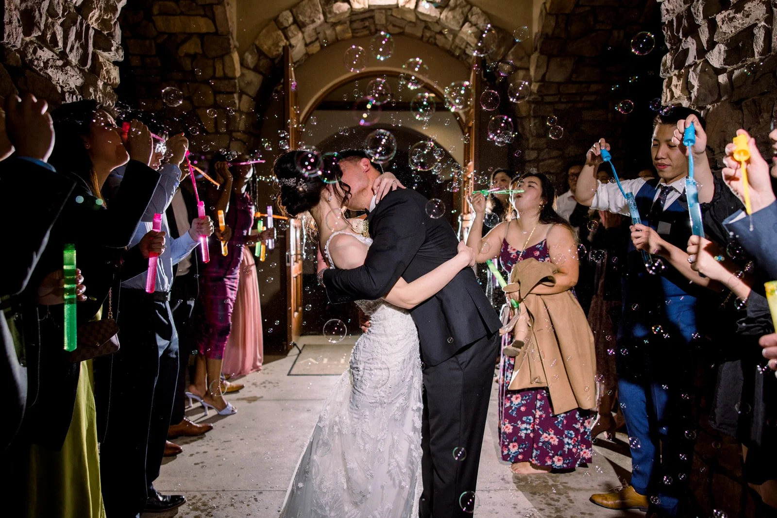 A bride and groom sharing a kiss surrounded by guests blowing bubbles during a wedding celebration.
