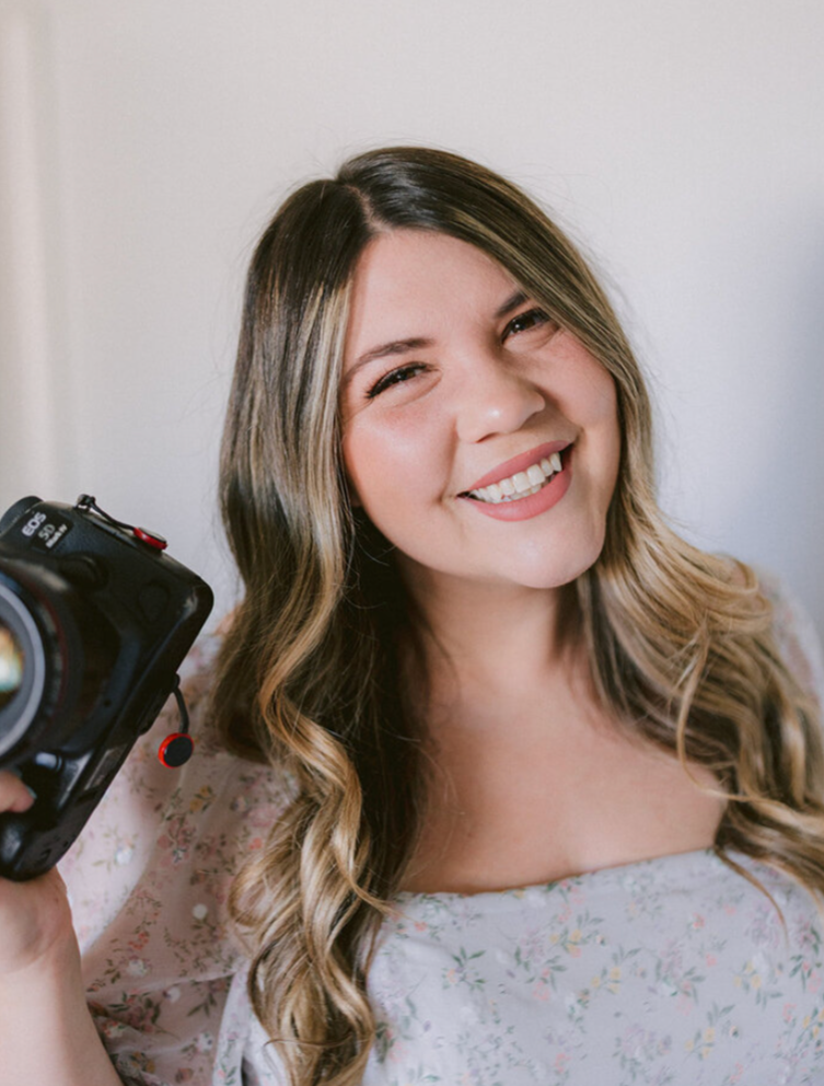 A woman with wavy blonde hair smiling and holding a camera.