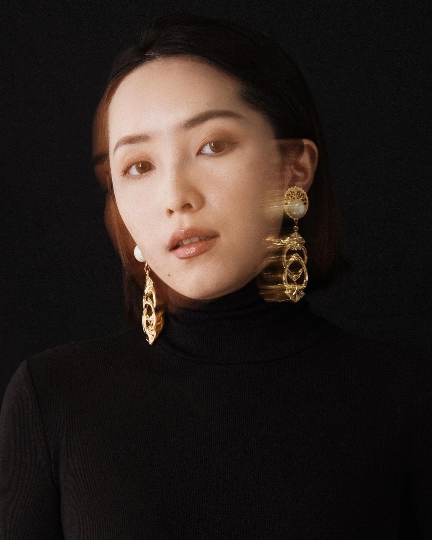 A woman wearing a black turtleneck and large gold earrings with a blurred motion effect on the right side of her face against a black background.
