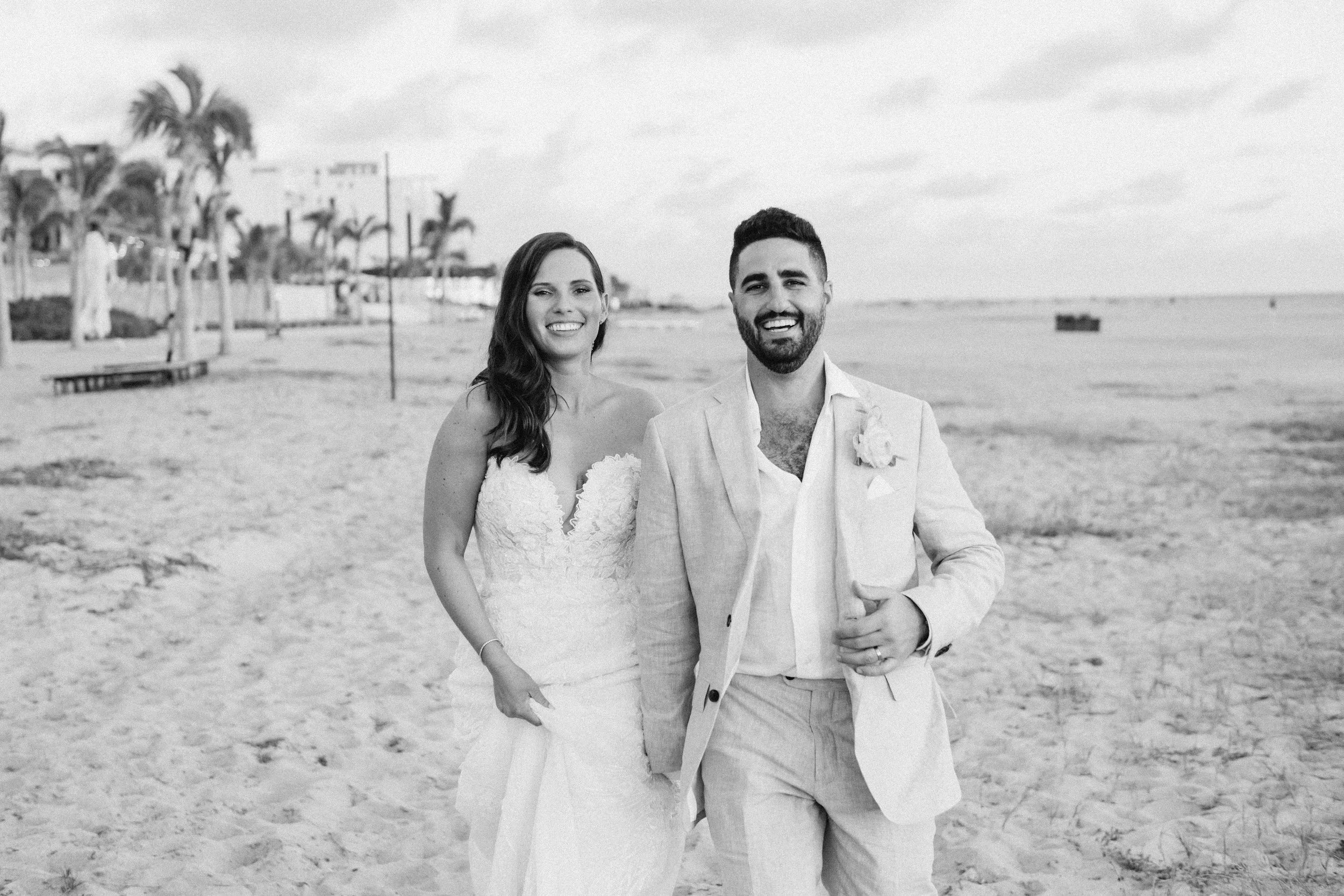 A smiling couple dressed in wedding attire walking on a beach, with palm trees and beachside houses in the background.