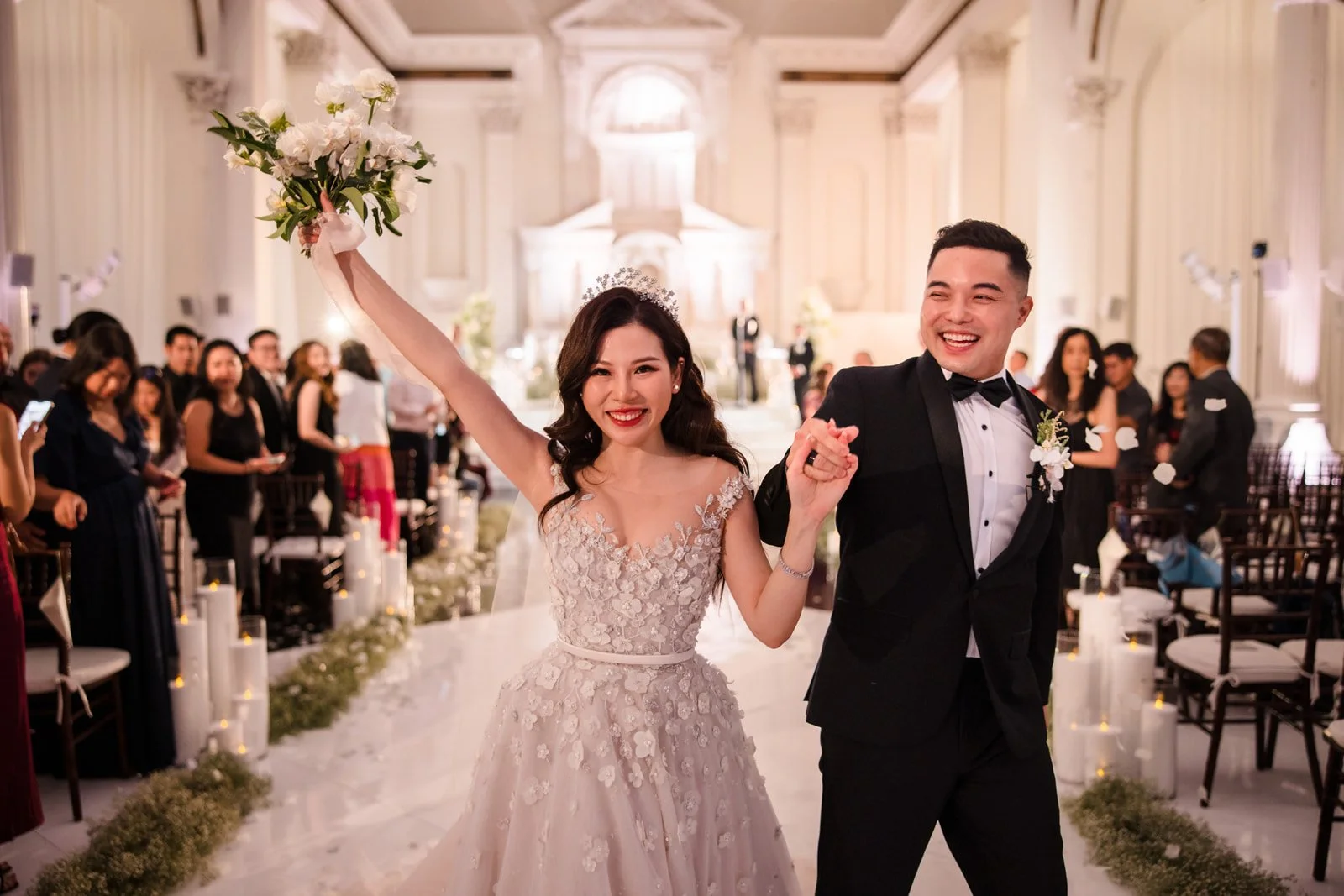 A newlywed couple celebrating their wedding in a grand hall. The bride is holding a bouquet of white flowers and wearing a lace wedding gown with floral appliqués and a tiara. The groom is wearing a black tuxedo with a bow tie. They are smiling and holding hands, surrounded by joyful guests.