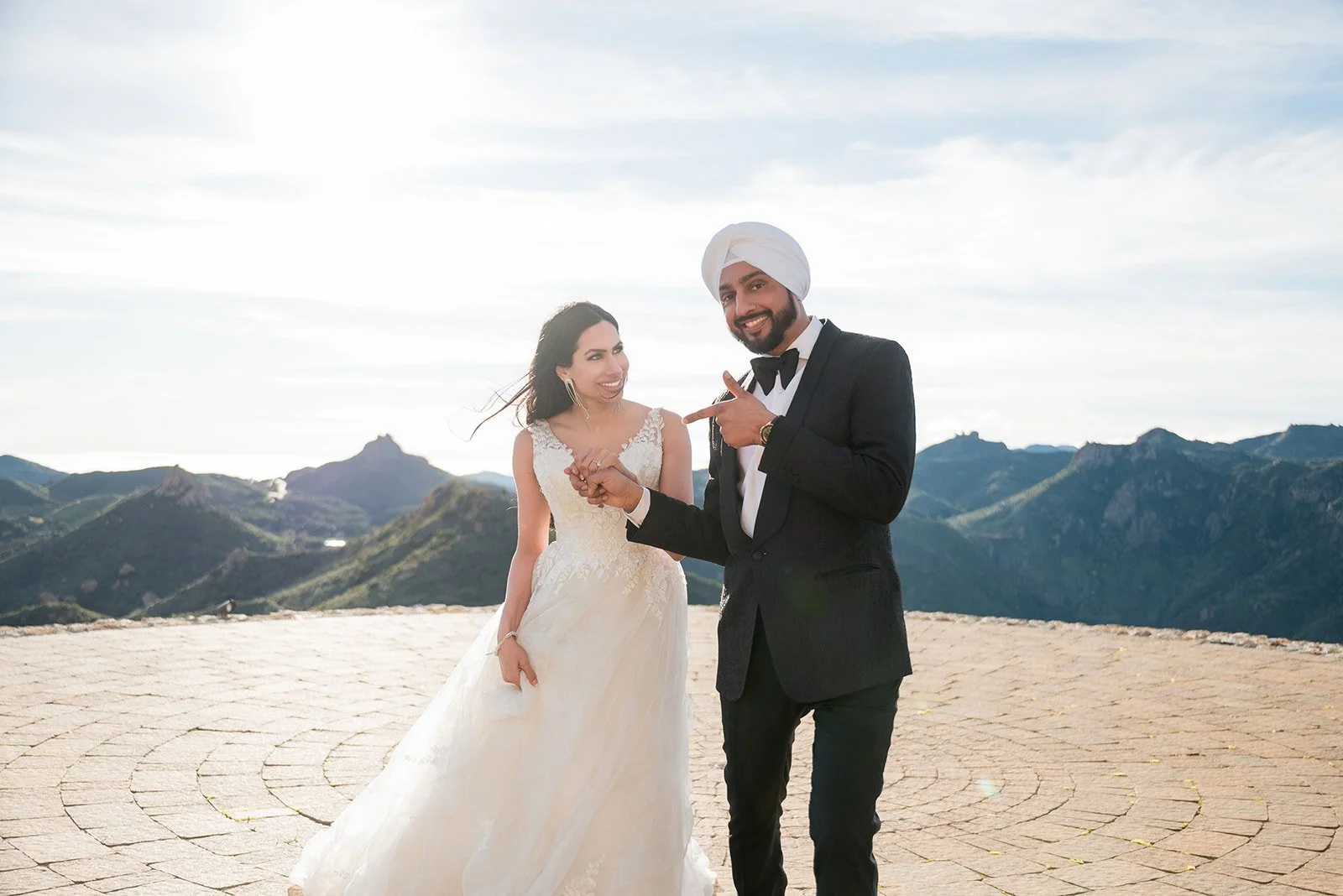 A newlywed couple dressed in wedding attire, standing outdoors with mountainous terrain in the background. The bride is wearing a white wedding gown, and the groom is wearing a black tuxedo with a white turban. They are smiling and holding hands, with the groom pointing at the bride.