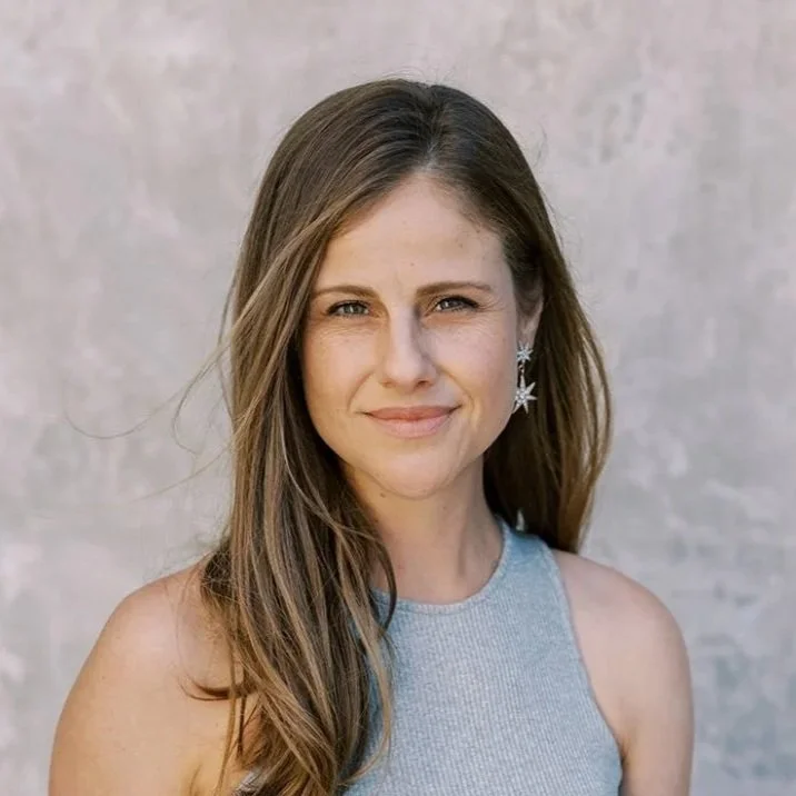 Portrait of a woman with long brown hair, wearing a light gray sleeveless top and star-shaped earrings, standing in front of a neutral background.