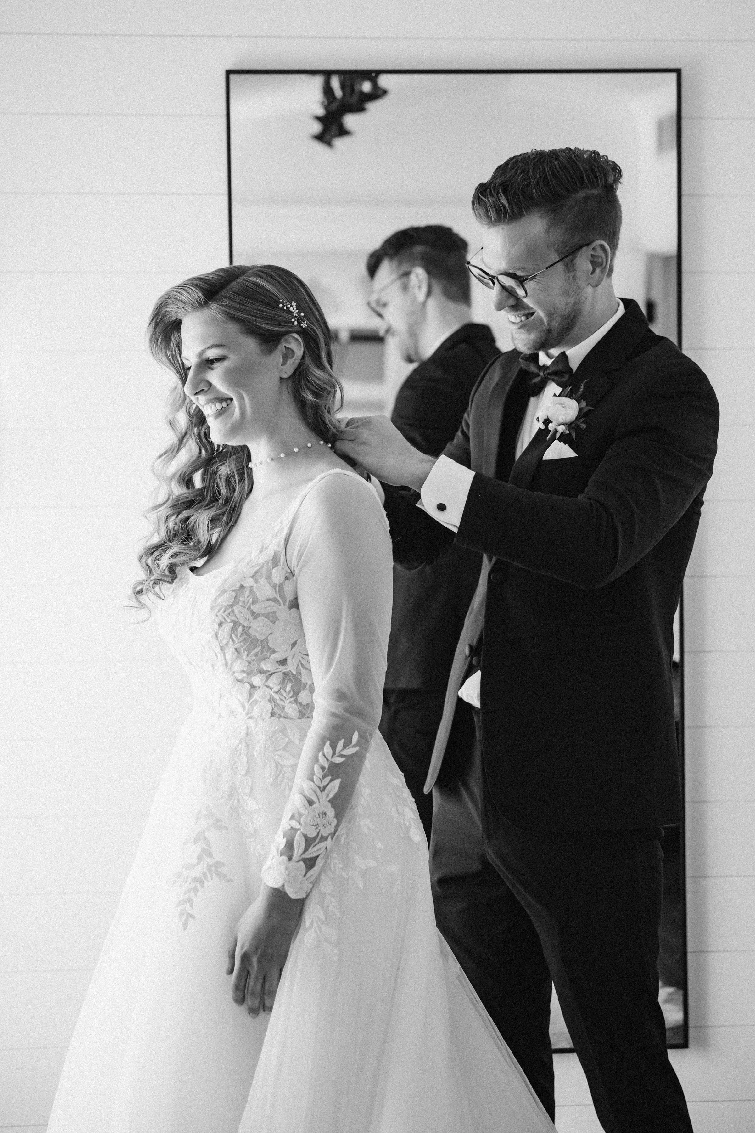 A bride with wavy hair and a floral wedding dress is smiling as a groom in a tuxedo helps secure her veil. In the background, a mirror reflects the couple and a man adjusting the bride's dress.