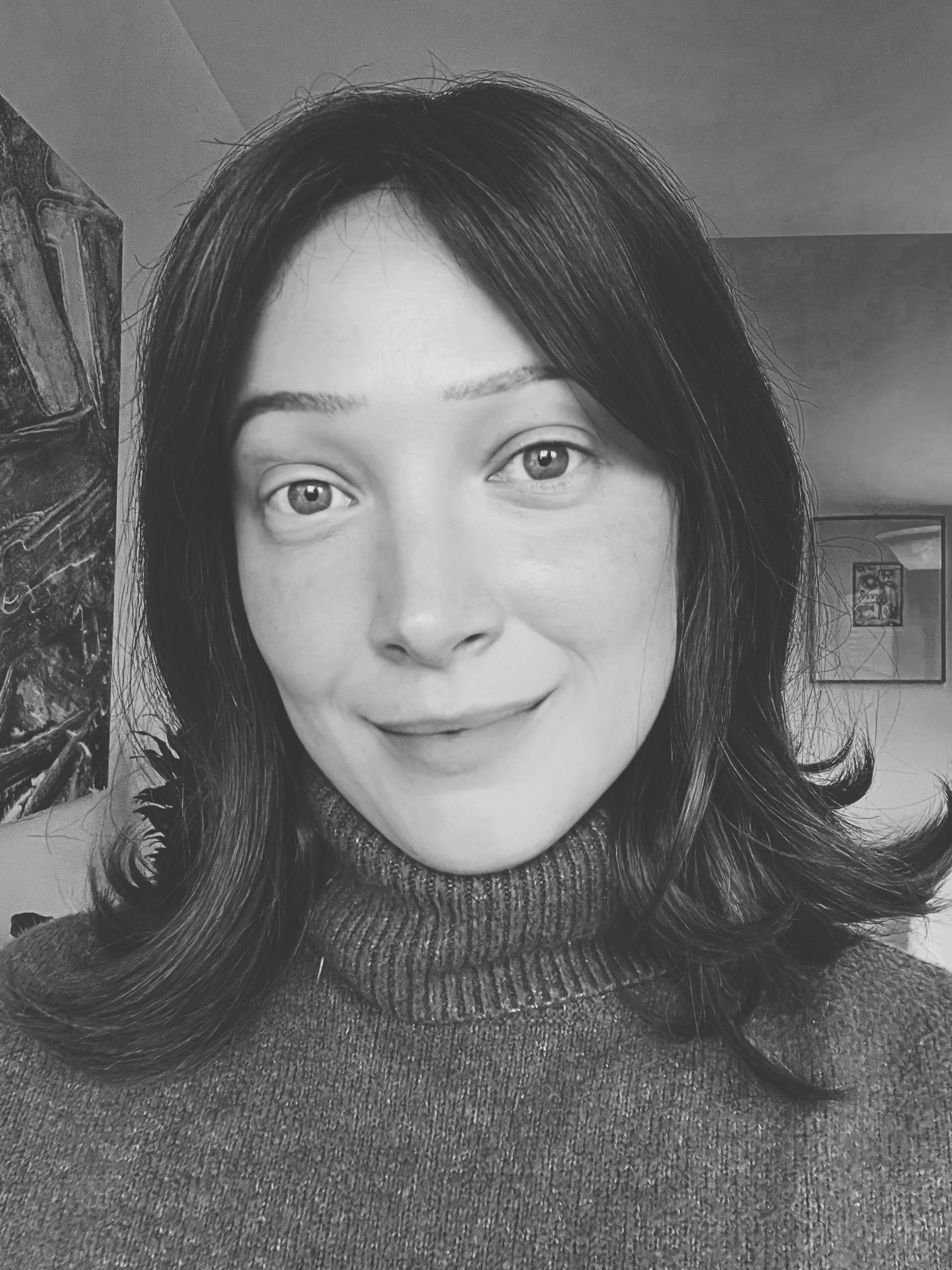 A black and white photograph of a woman, Erin McQueen, with shoulder-length dark hair, wearing a turtleneck sweater, smiling softly at the camera in an indoor setting.