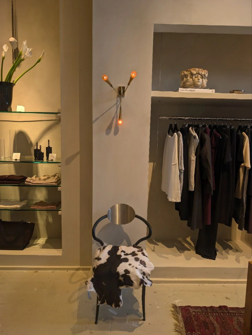 Interior of a clothing store with shelves of folded clothes, a rack of hanging garments, a modern wall lamp, a decorative stone sculpture, and a chair covered with a cowhide rug.