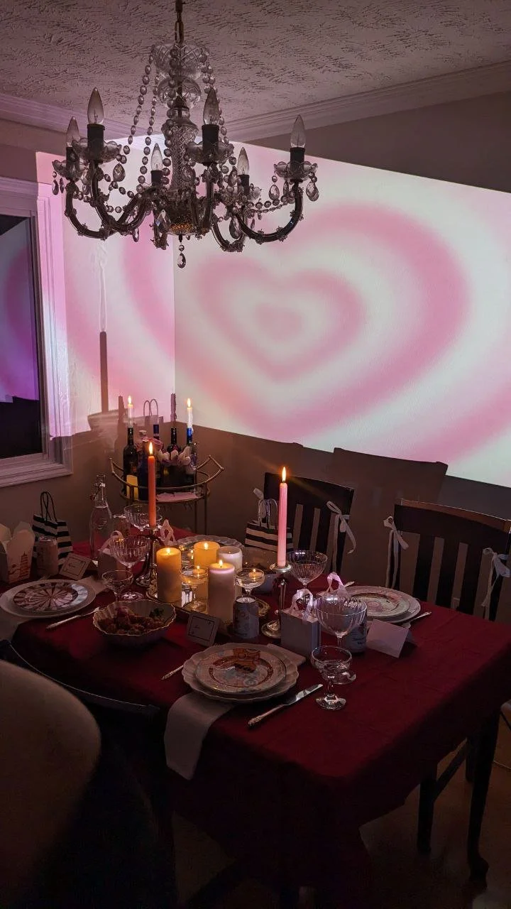 A dining table set for a romantic dinner with candles, wine, and decorative plates, illuminated by a chandelier and pink heart-shaped projection on the wall.