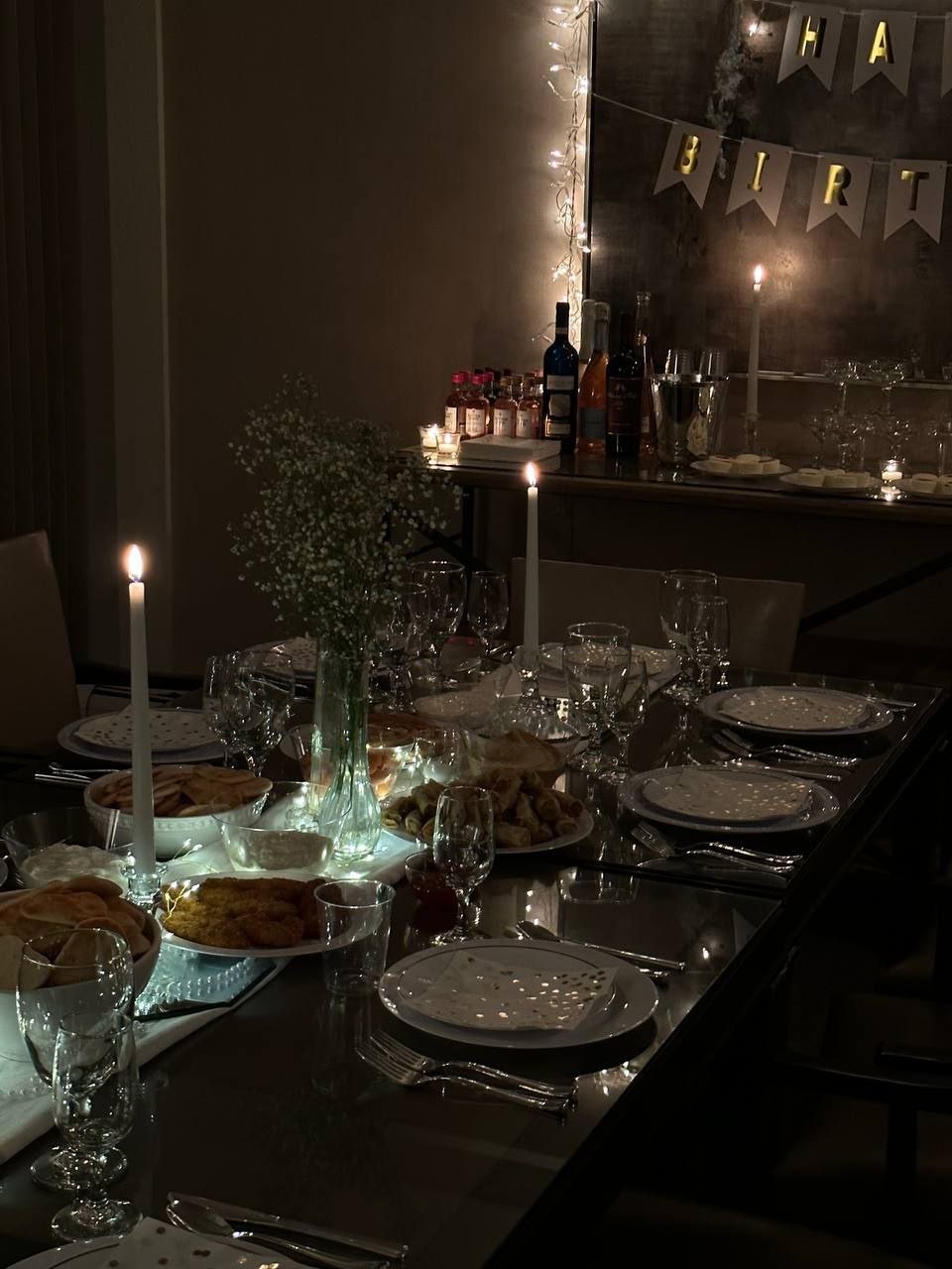A dimly lit dining table set for a celebration with plates, glasses, candles, and a floral centerpiece, with a 'Happy Birthday' banner in the background.
