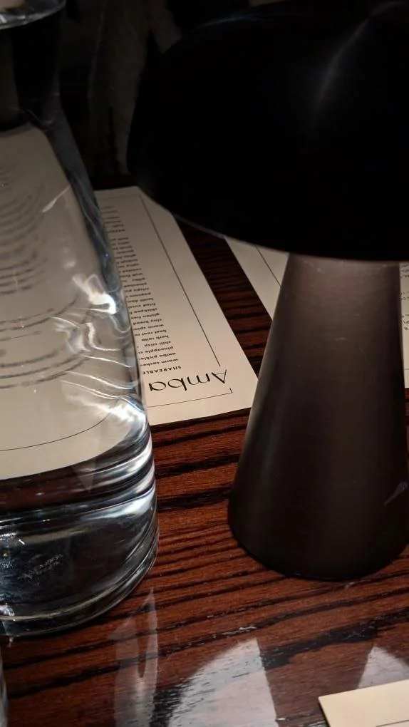 Close-up of a clear glass water bottle, a black table lamp, and a restaurant menu on a dark wooden table.