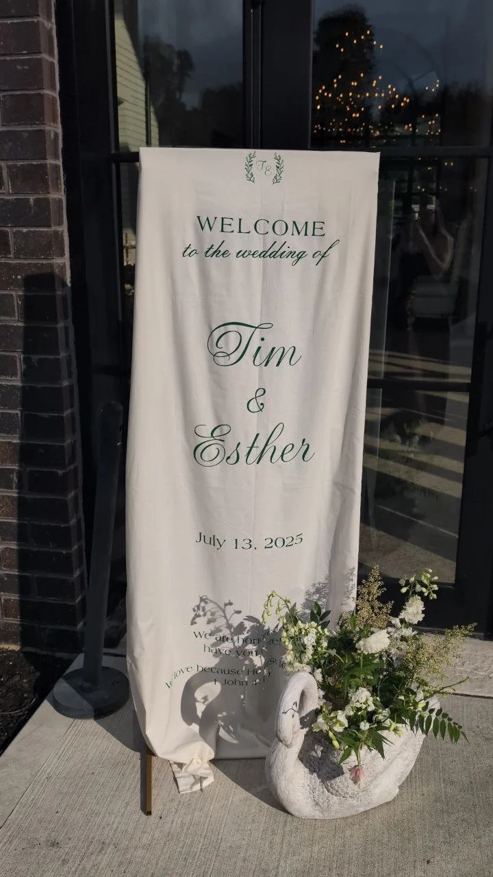 A welcome sign for a wedding on July 13, 2025, with the names Tim and Esther. The sign is white with green text and decorative elements, and it is placed next to a flower arrangement in a stone swan-shaped planter outside a building.