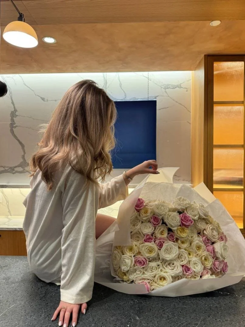 A woman with wavy blonde hair sitting at a kitchen counter, holding a large bouquet of white and pink roses.