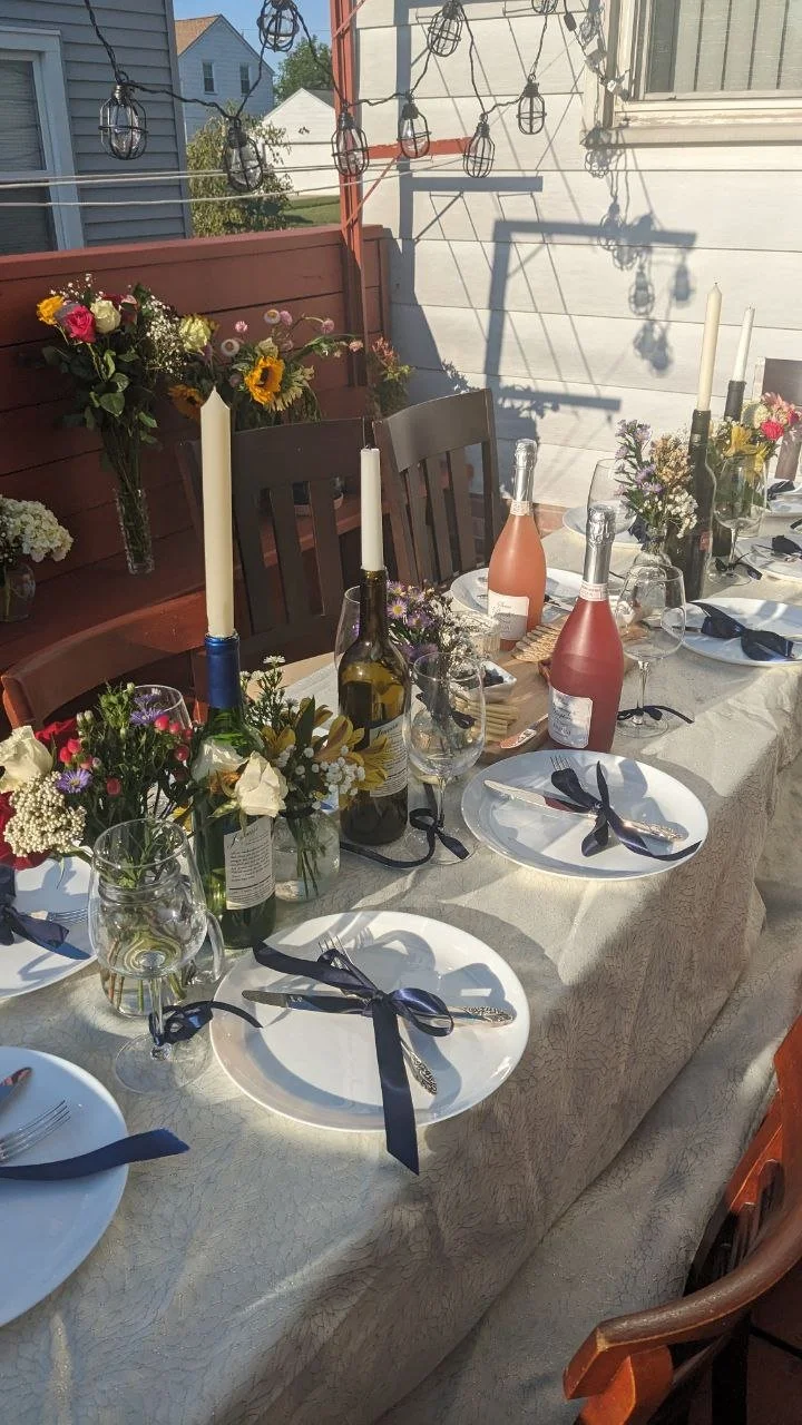 Outdoor dining table set for a celebration with floral centerpieces, bottles of rosé wine, candles, and black utensils tied with navy ribbons, on a sunlit patio with string lights overhead.