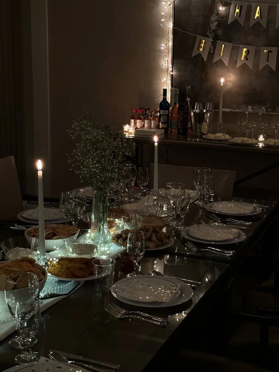 A dimly lit dining room decorated for a birthday celebration with a 'Happy Birthday' banner, a table set with plates, glasses, and candles, and a side table with bottles of alcohol, glasses, and snacks.