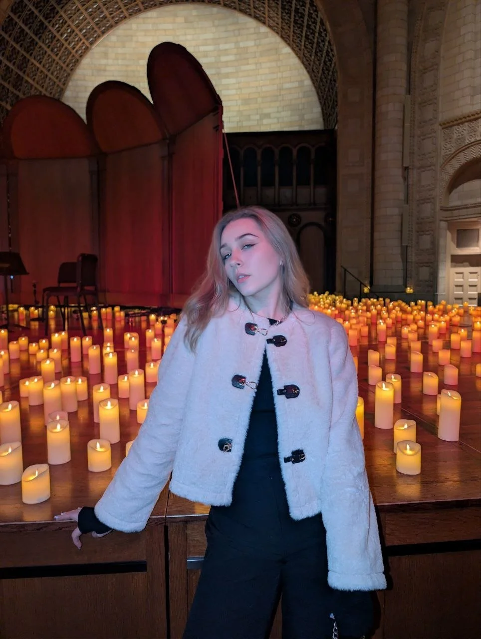 A young woman with long blonde hair posing inside a church or cathedral, surrounded by lit candles on a wooden surface, with arched architectural features and a wooden partition in the background.