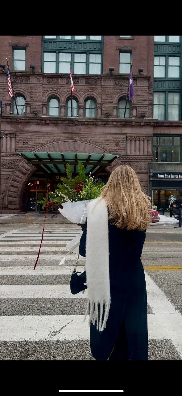 A woman with long blonde hair carrying a flower bouquet crosses a city street with a large brownstone building in the background. She is wearing a dark coat and a white scarf.