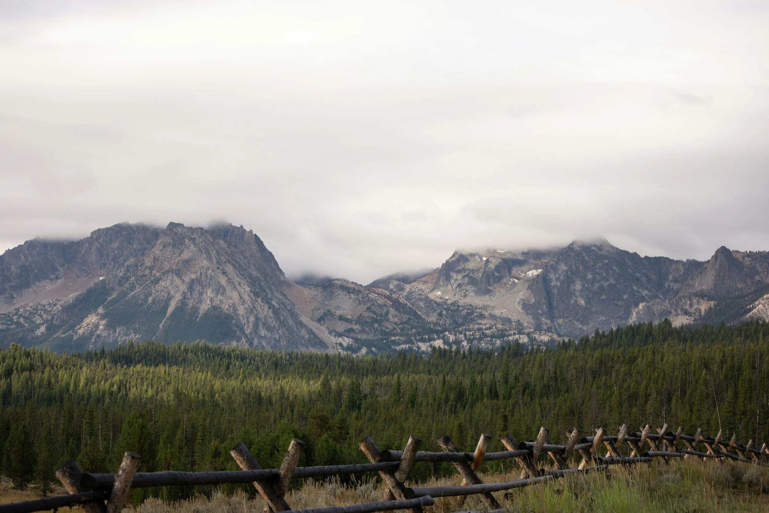 A mountain range with peaks partially covered by clouds, dense green forest below, and a rustic wooden fence in the foreground.