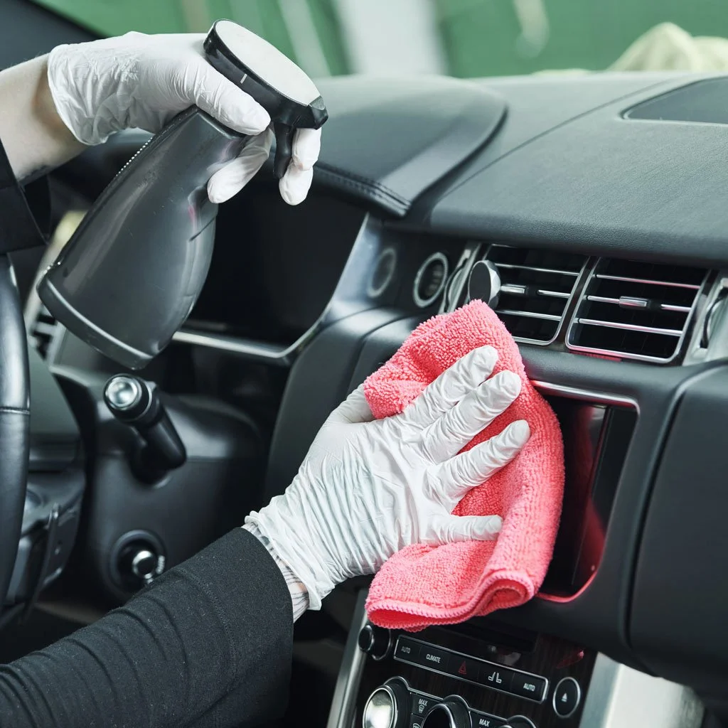 Person cleaning a car's dashboard with a spray bottle and a pink microfiber cloth, wearing white gloves.