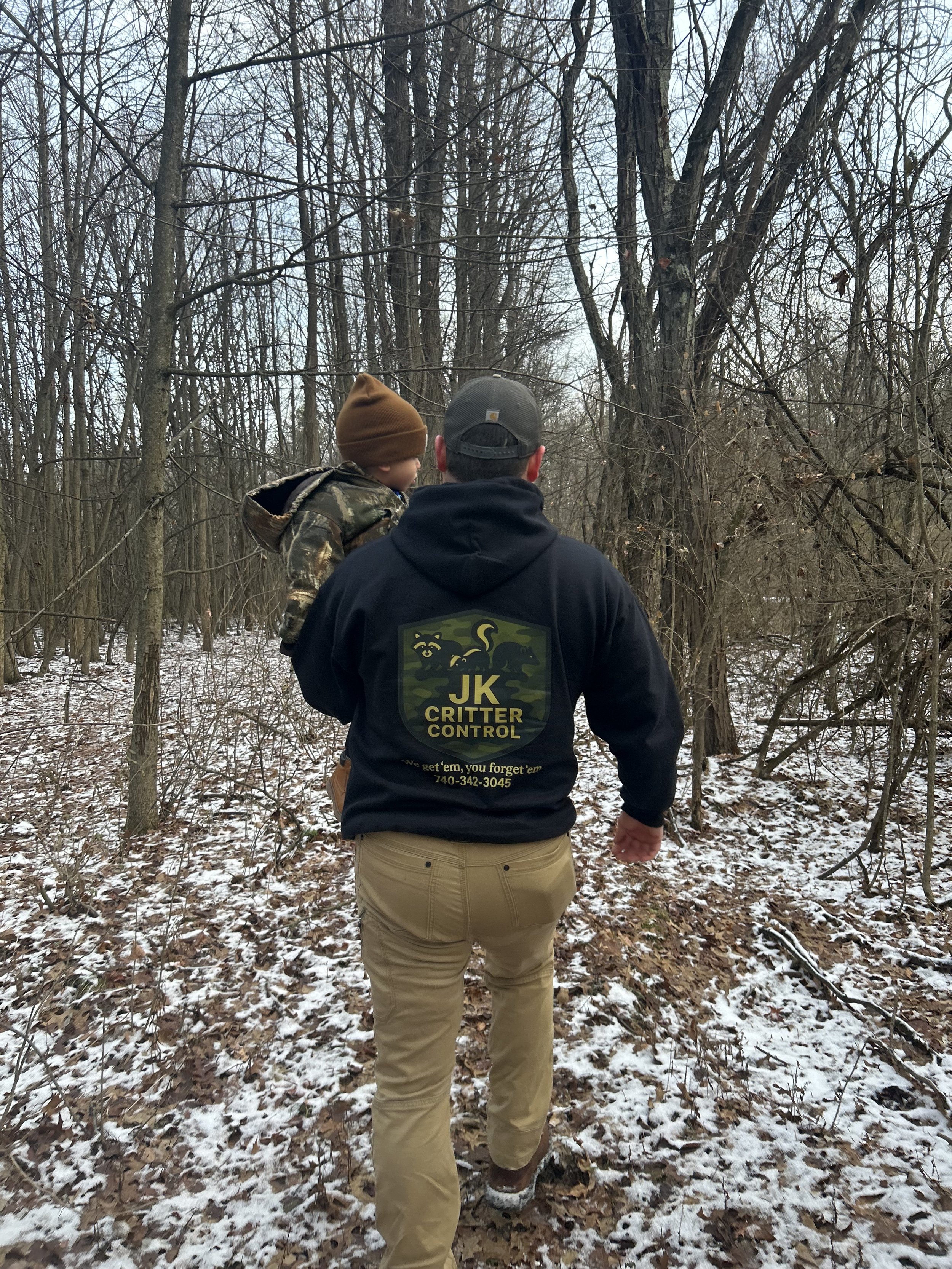 A man carrying a young boy through a wooded area in winter. The man is wearing a black hoodie with a logo and text that reads 'JK CRITTER CONTROL,' and khaki pants. The boy is dressed in a camouflage jacket and brown hat. The ground is covered with snow and fallen leaves, and bare trees surround them.
