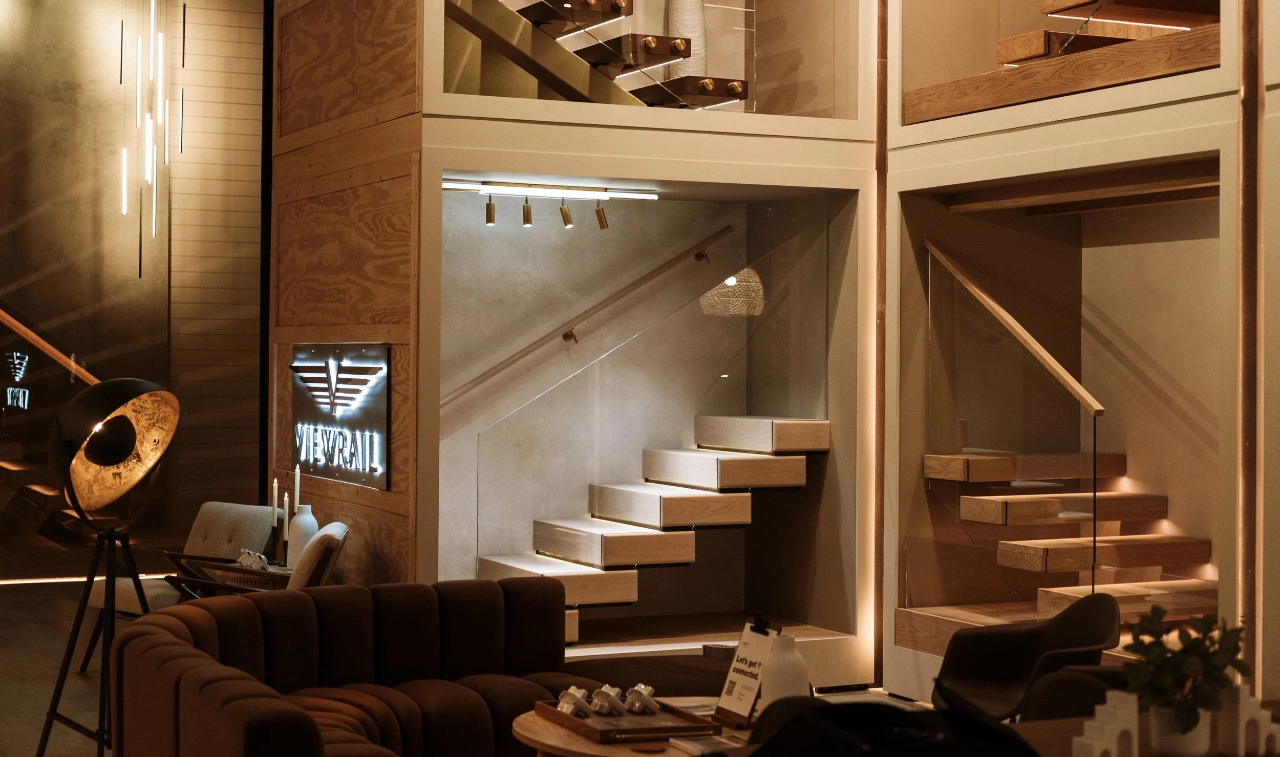 Interior of a modern furniture store showcasing spiral staircases with wooden steps and glass railings, stylish lighting, a sofa, a floor lamp, and a neon sign reading 'VIEW RAIN'.