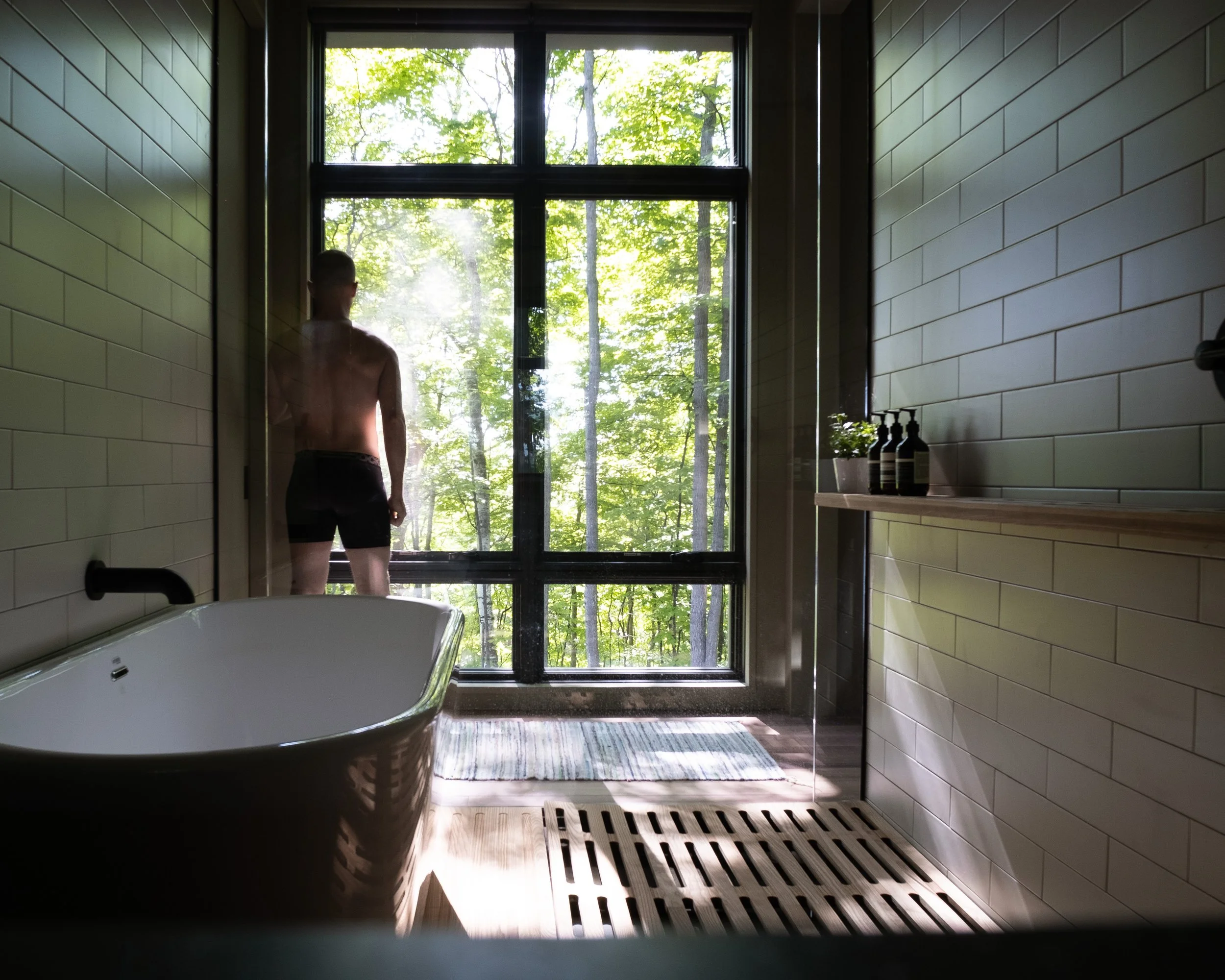 A person standing partially covered in steam, facing a large window with a view of green trees outside, in a bathroom with a bathtub, beige tiled walls, and bottles on a shelf.