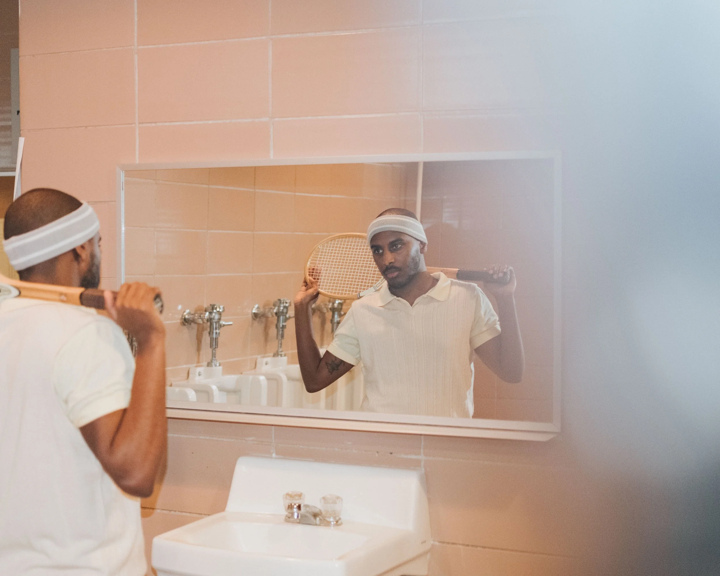 A man with a tennis racket and headband looking at himself in a bathroom mirror, with sink and beige tiled wall.