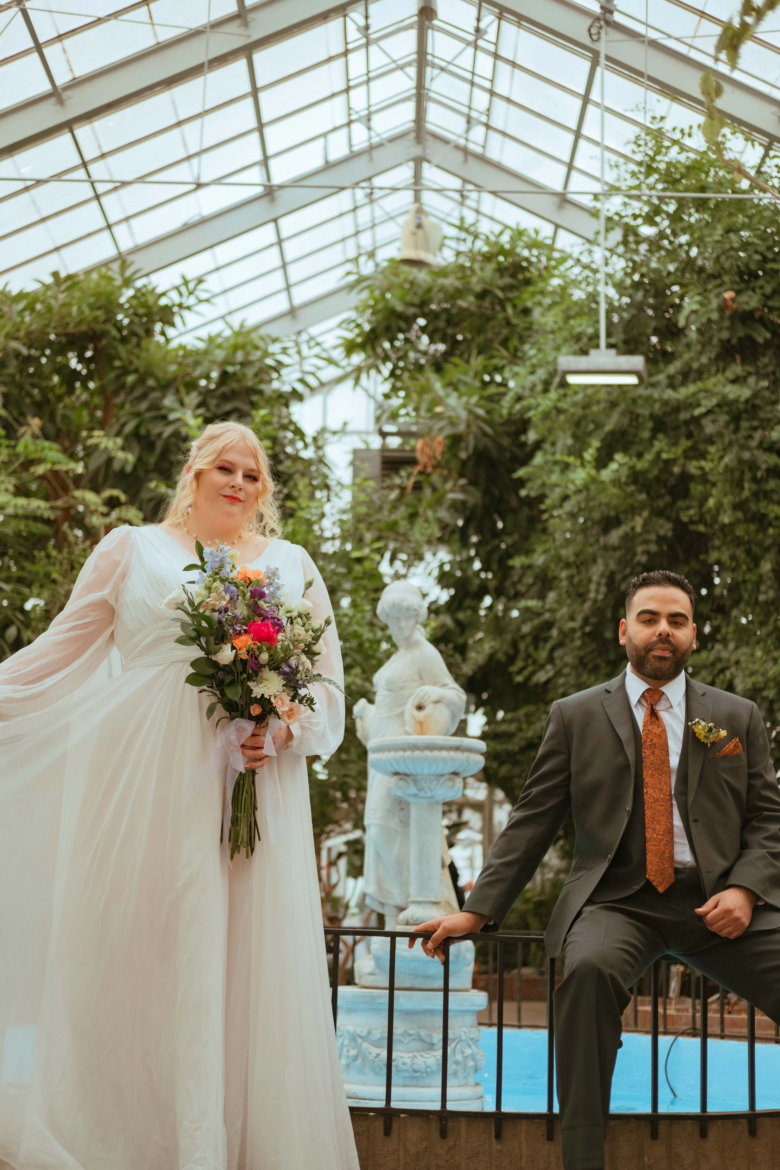 A bride in a white dress holding a colorful bouquet and a groom in a dark suit with an orange tie, posing together in a greenhouse setting surrounded by green plants and a decorative white statue.