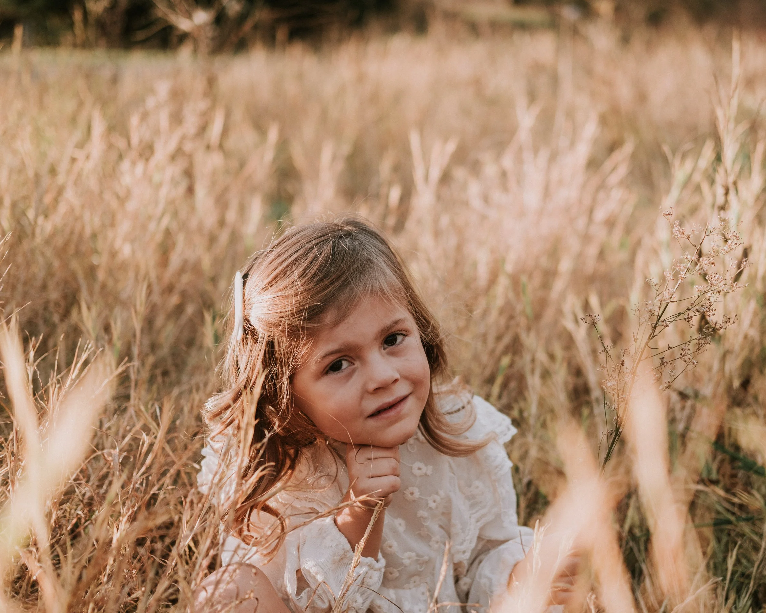 A young girl with light brown hair sitting in a field of tall, golden grass during golden hour, wearing a white lacy dress, looking at the camera with a slight smile, holding her chin with one hand.