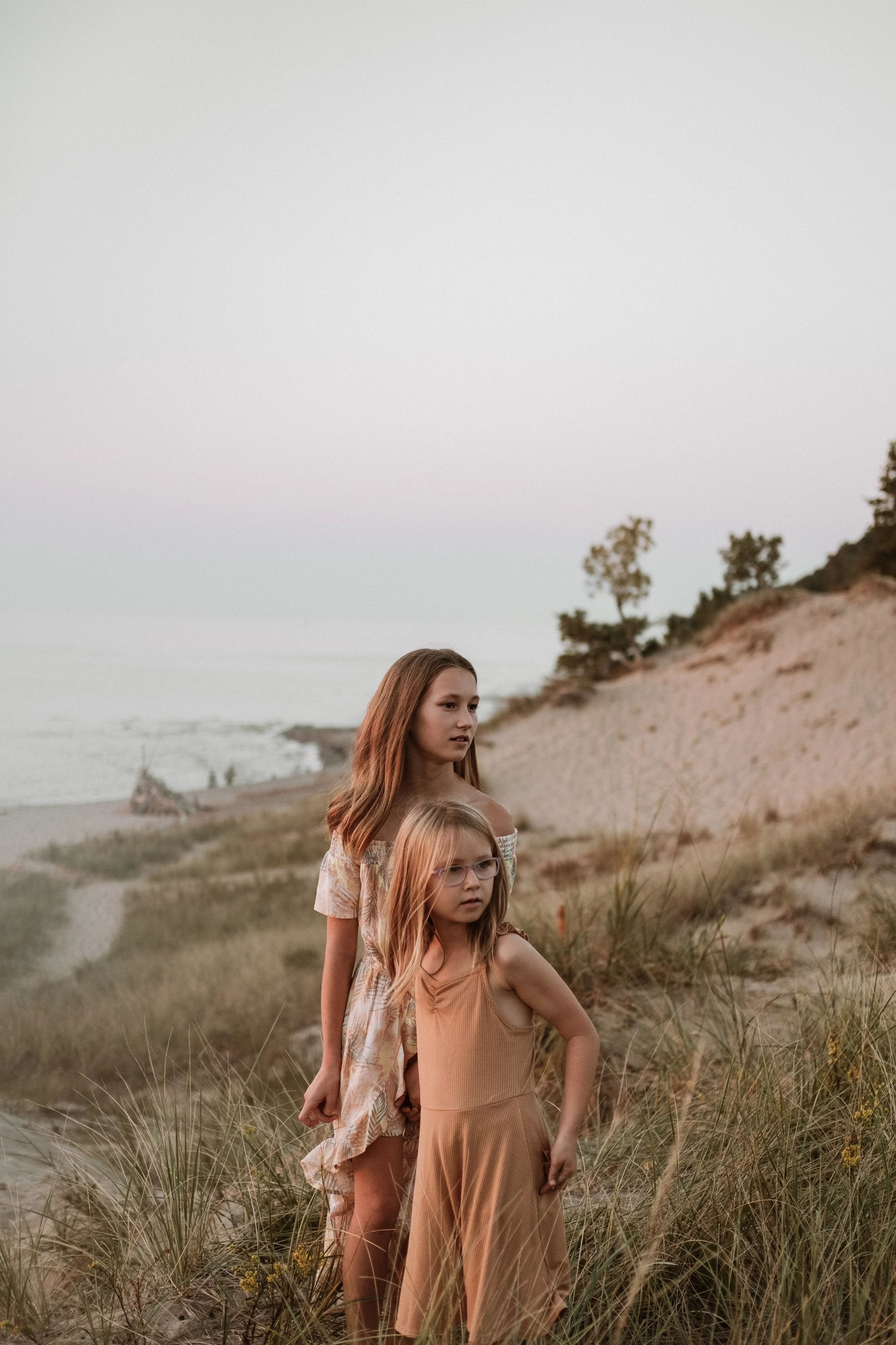 Two young girls standing on a grassy beach dune, looking thoughtfully into the distance with ocean and sky in the background during sunset.