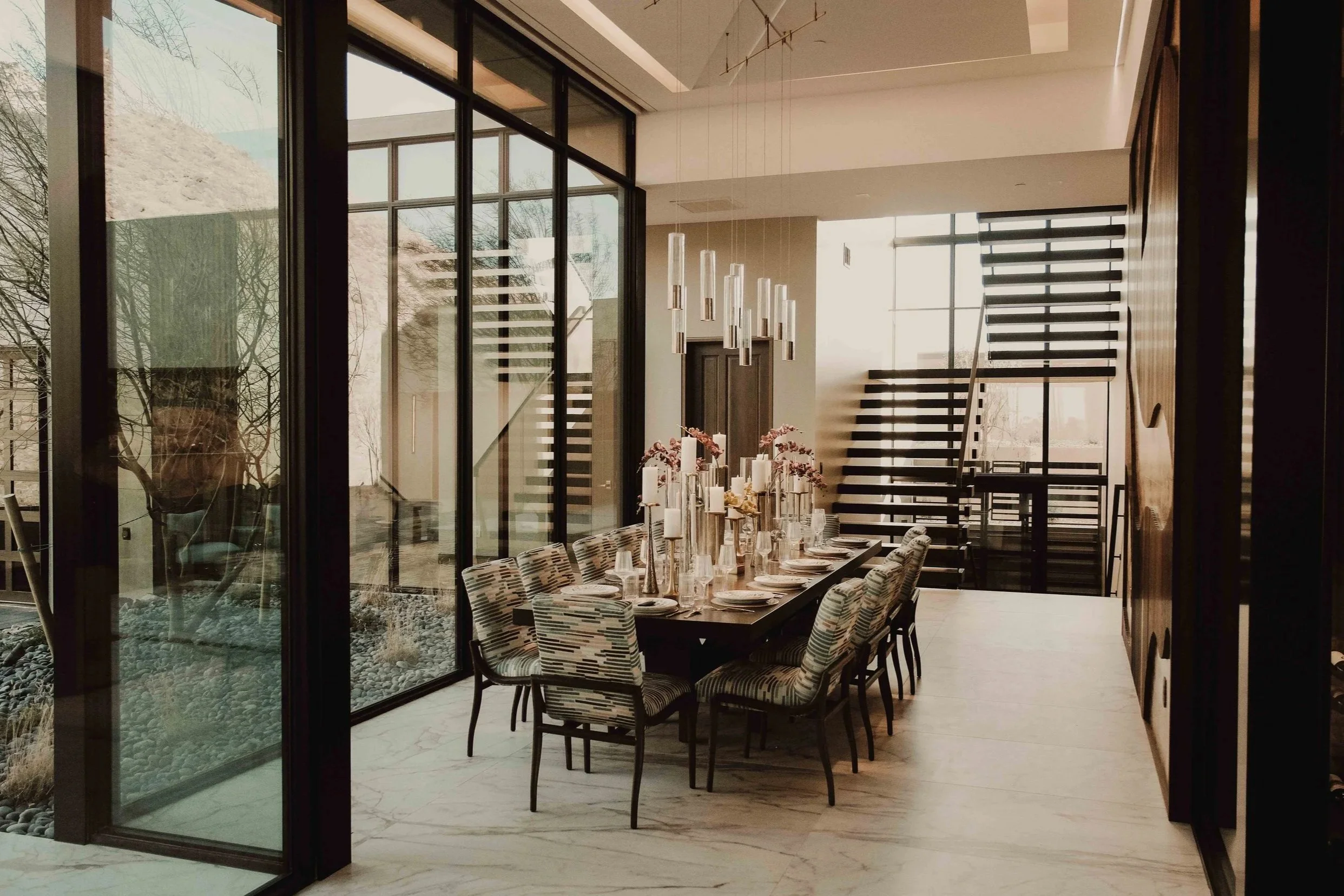 Modern dining room with a long table set with plates, glasses, and pink floral centerpieces, surrounded by patterned chairs, next to large glass walls and a staircase with open risers.