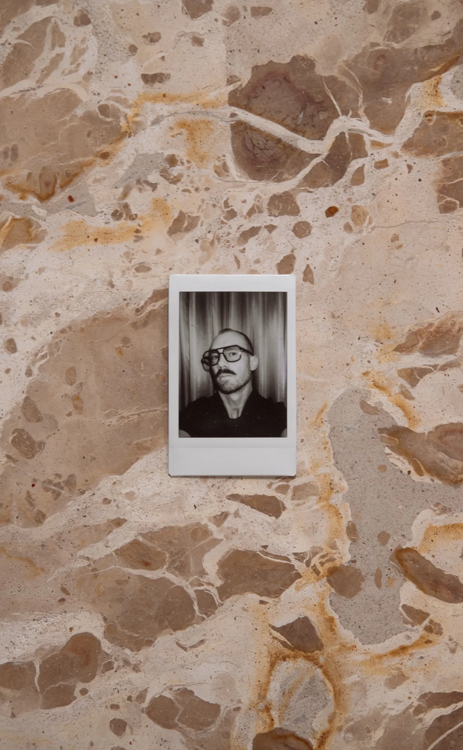 Black and white photograph of a man with a beard and glasses, standing in front of curtains, with one eye partially covered by a sheer fabric.