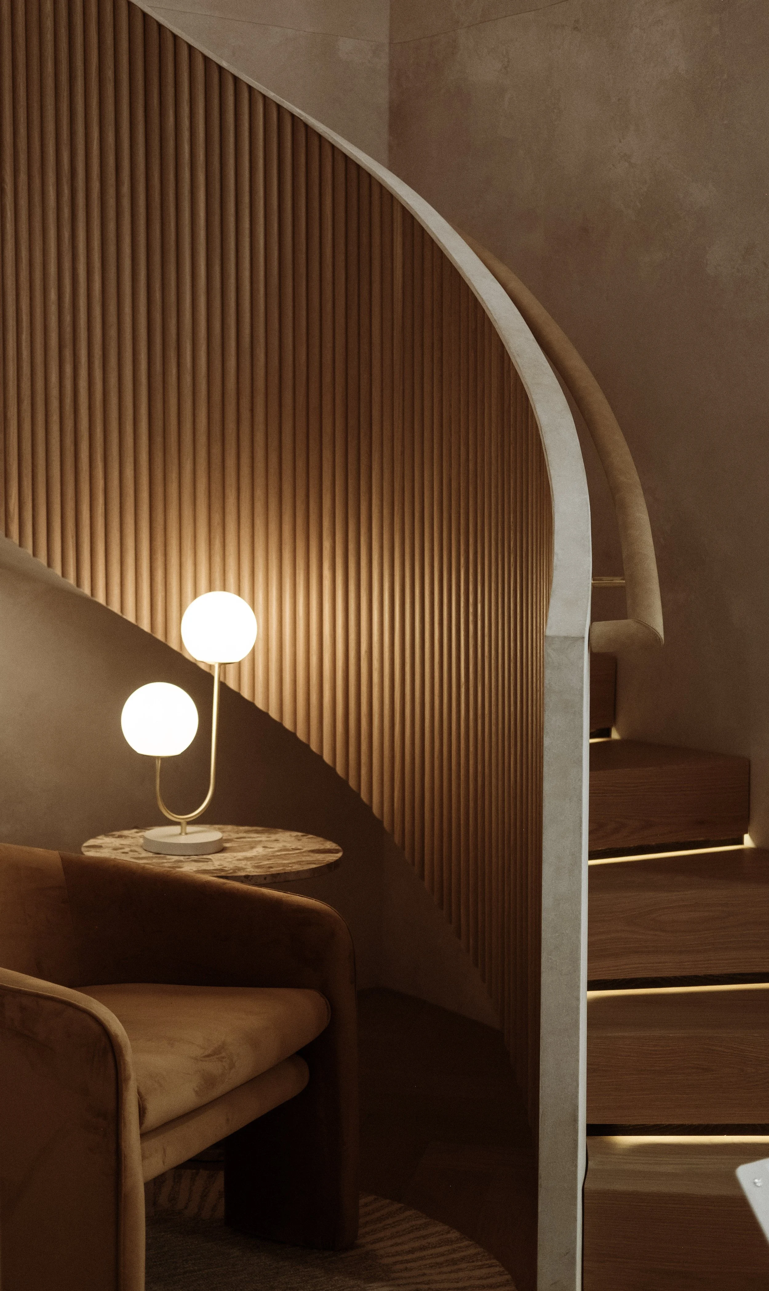 Contemporary interior with a curved wooden partition, a beige armchair, a marble-topped side table, and a modern lamp with two round white bulbs.