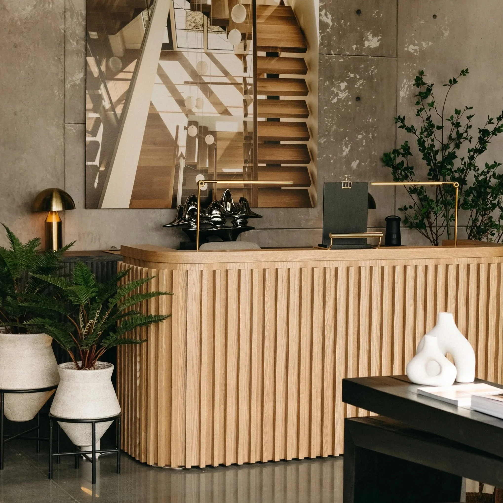 Modern reception desk with wooden slats, a green plant in a white pot, and decorative objects including a black sculpture, a white abstract sculpture, and a black book in a stylish interior setting.