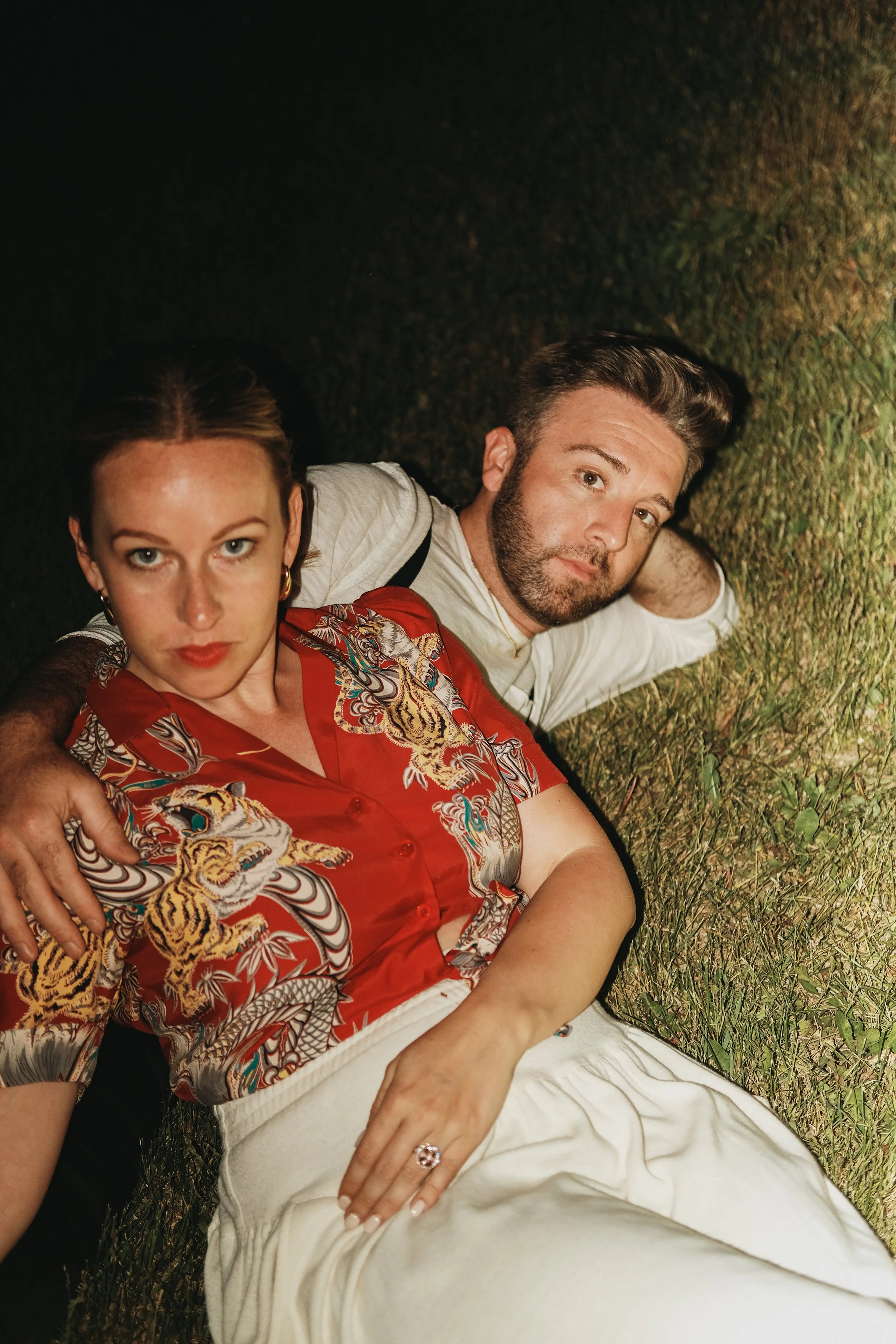 A man and woman lying on the grass at night, looking at the camera. The woman is in the foreground wearing a red patterned shirt, and the man is behind her wearing a white shirt.