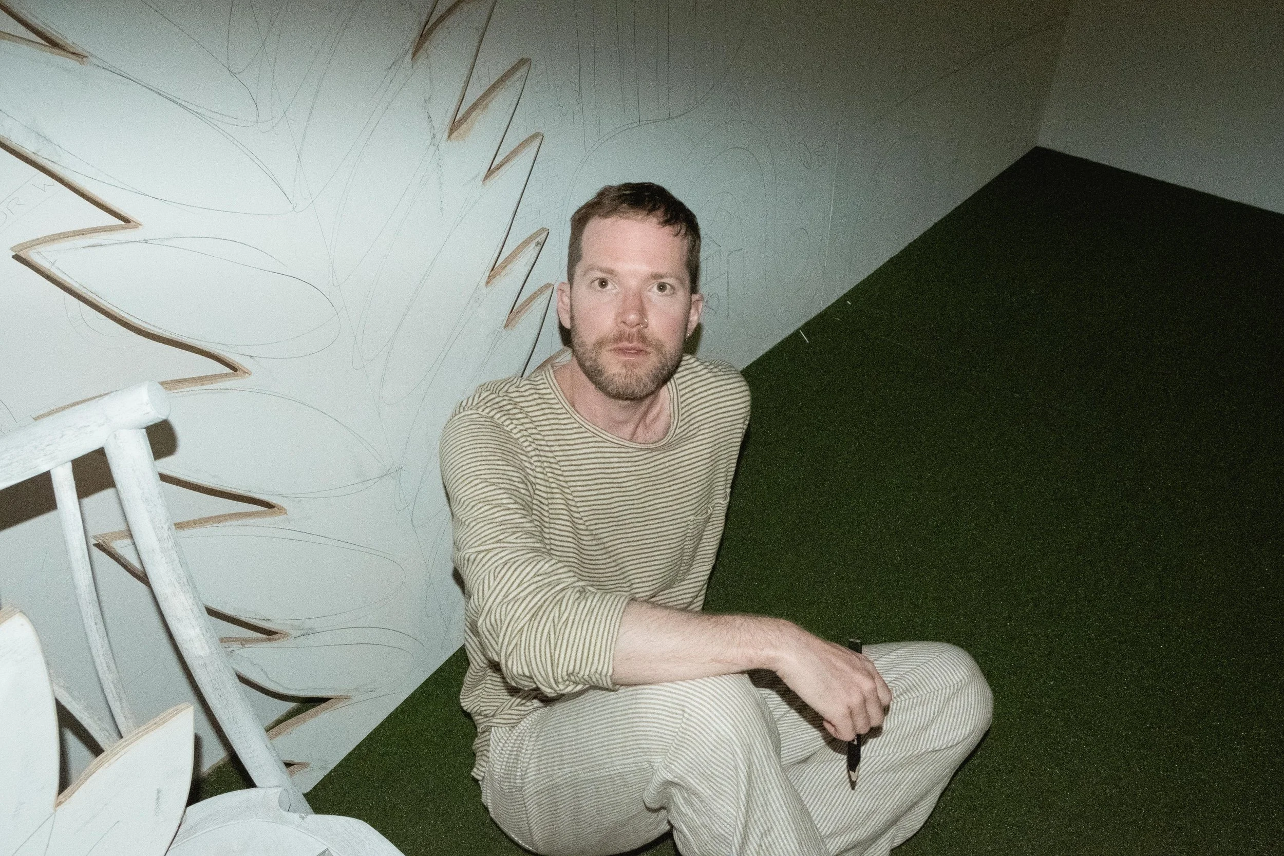 A man with a beard and short brown hair sitting on a green carpeted floor, looking up at the camera, wearing a striped shirt and light-colored pants. The background features a wall with abstract black line drawings and some white, leaf-shaped wooden 
