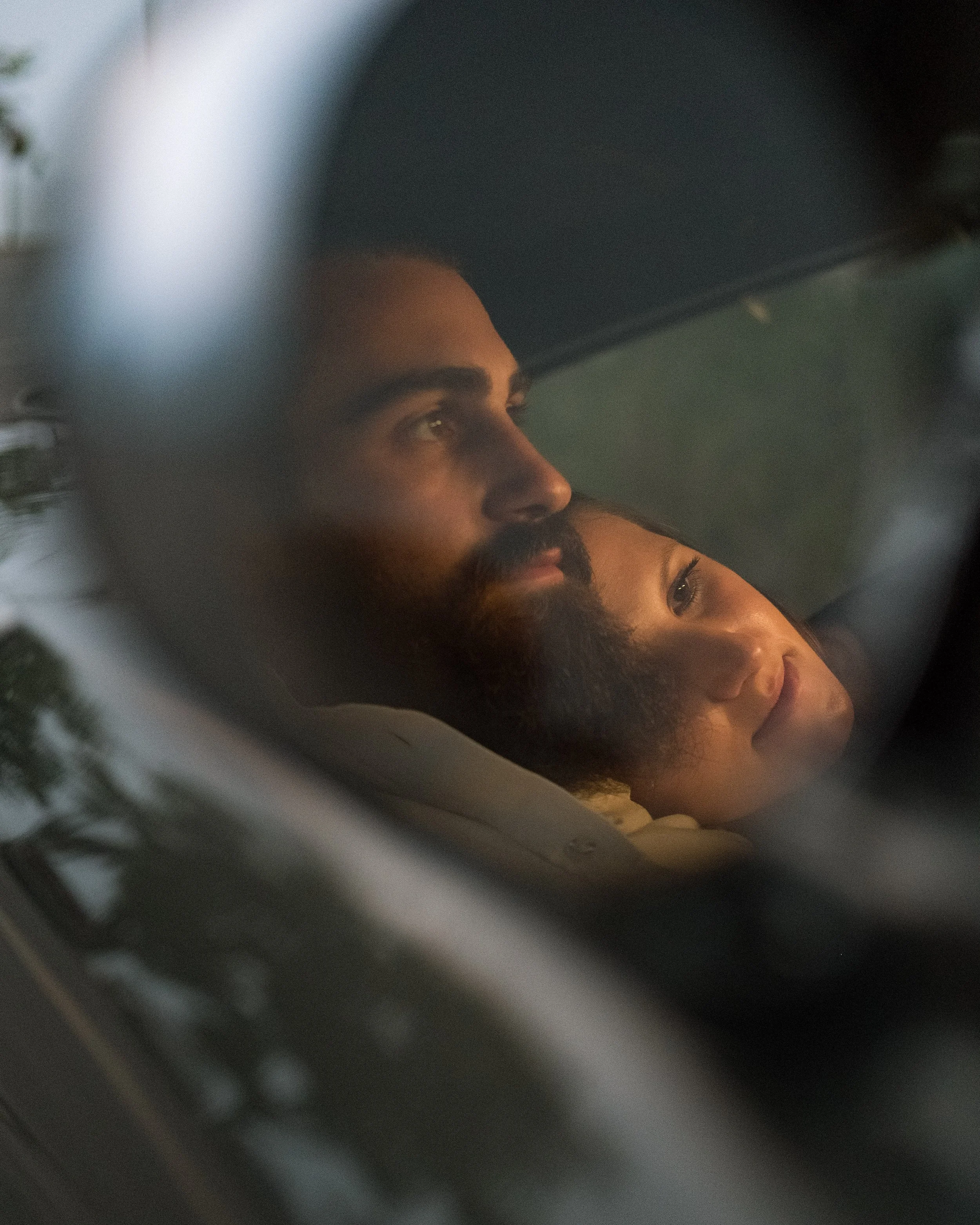 A close-up photo of a couple, viewed through a car window, lying down inside the vehicle, looking out contemplatively.